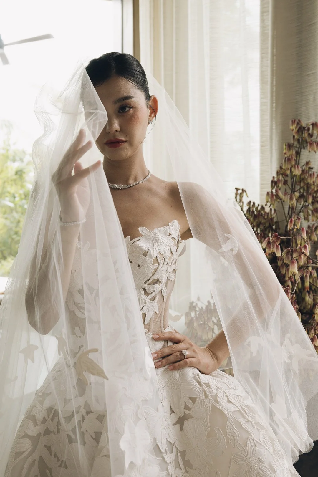 A bride in a white wedding gown with floral lace details, standing indoors near a window, holding her veil with one hand, and showing her engagement ring with the other, with plants in the background.