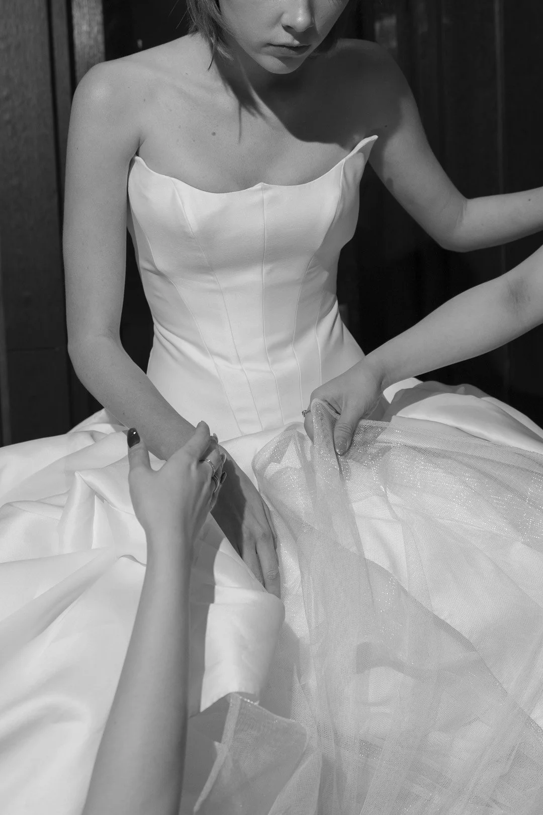 A woman in a strapless wedding dress is getting dressed, with another person's hands helping to adjust the gown and tulle fabric.