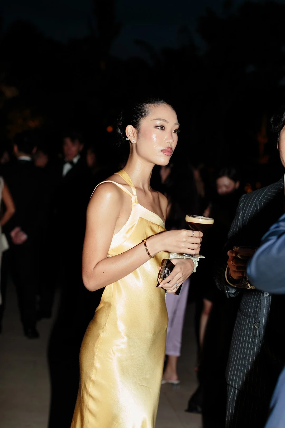 A woman with dark hair styled back, wearing a shiny gold camisole dress, holding a cocktail glass, standing at a social event at night with people in formal attire in the background.
