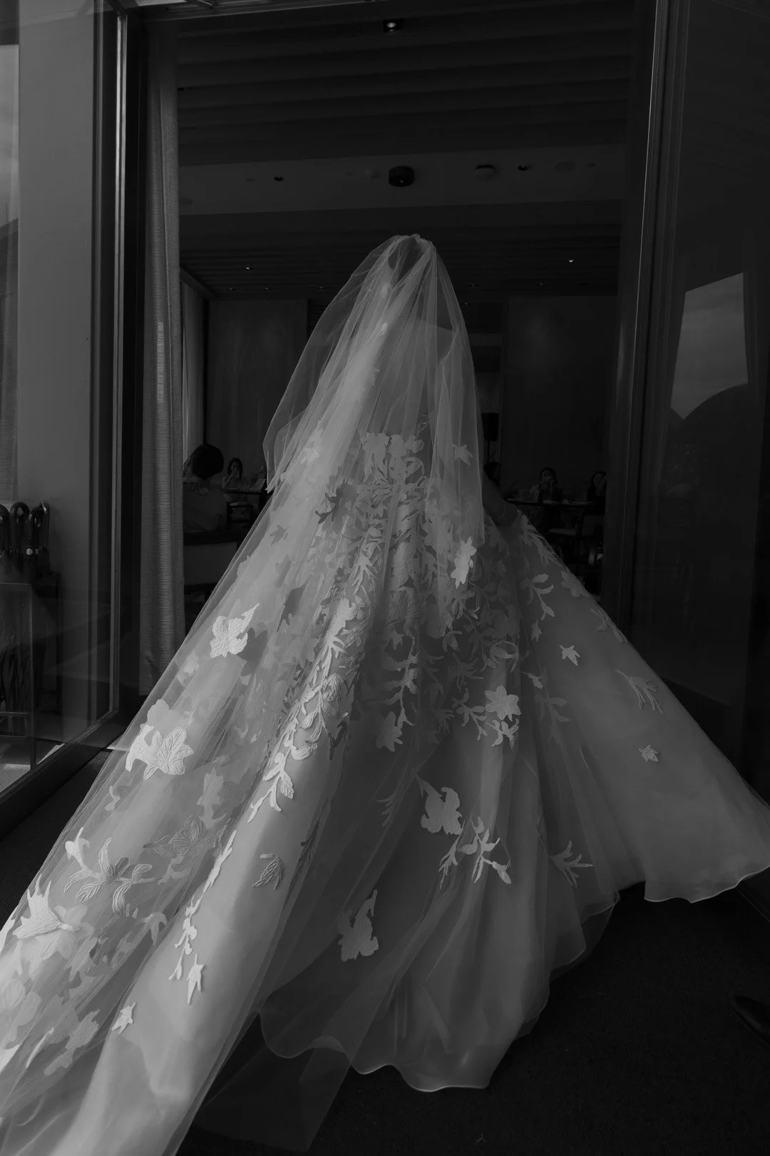 A wedding dress with floral lace details hanging inside a room, viewed through a glass door with reflection of people sitting at tables.