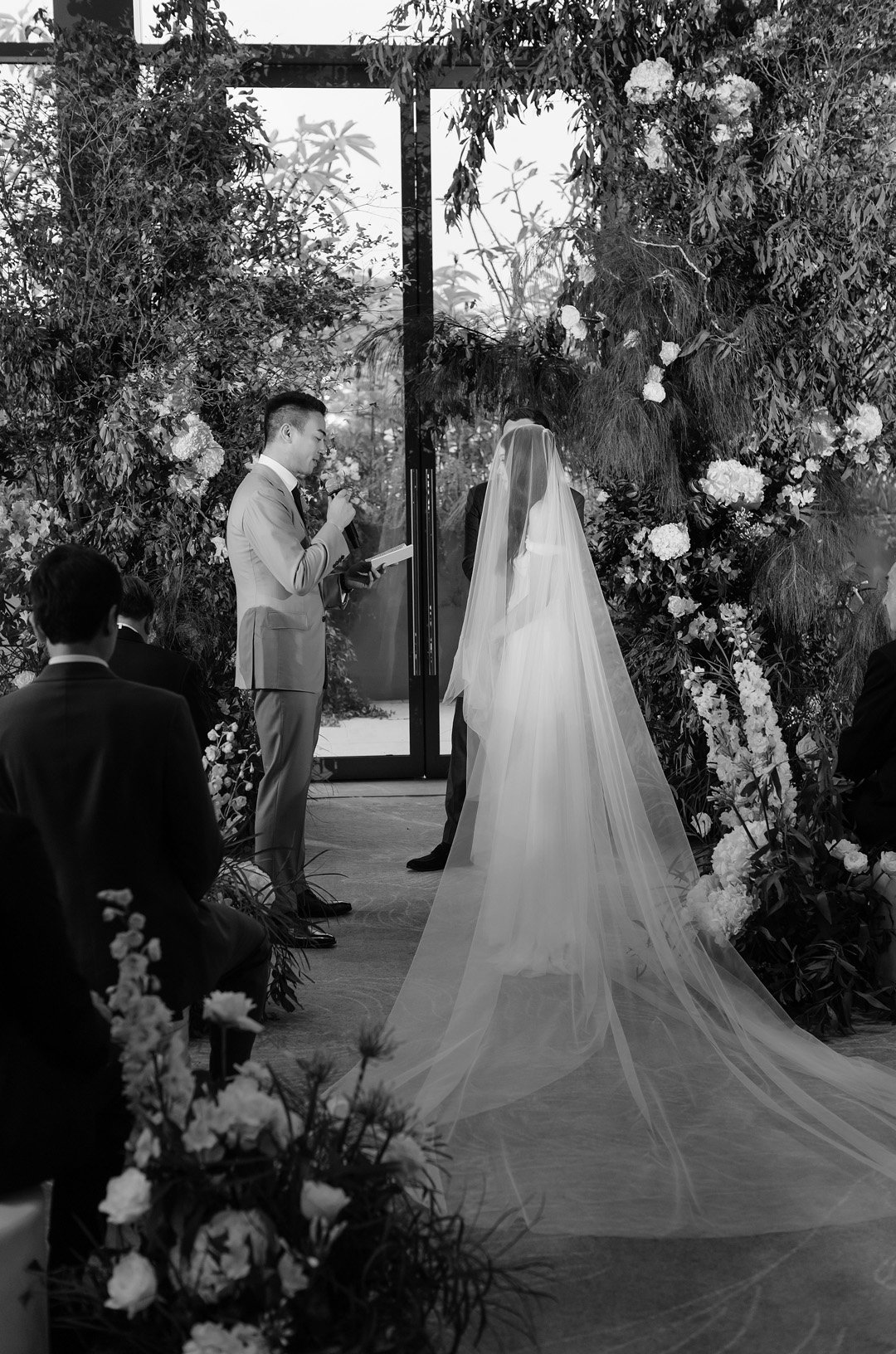 A black-and-white photo of a wedding ceremony with a bride in a white dress and veil, standing beside a groom in a suit. An officiant is reading from a book. The setting is decorated with flowers and greenery, and there is a glass door in the backgro