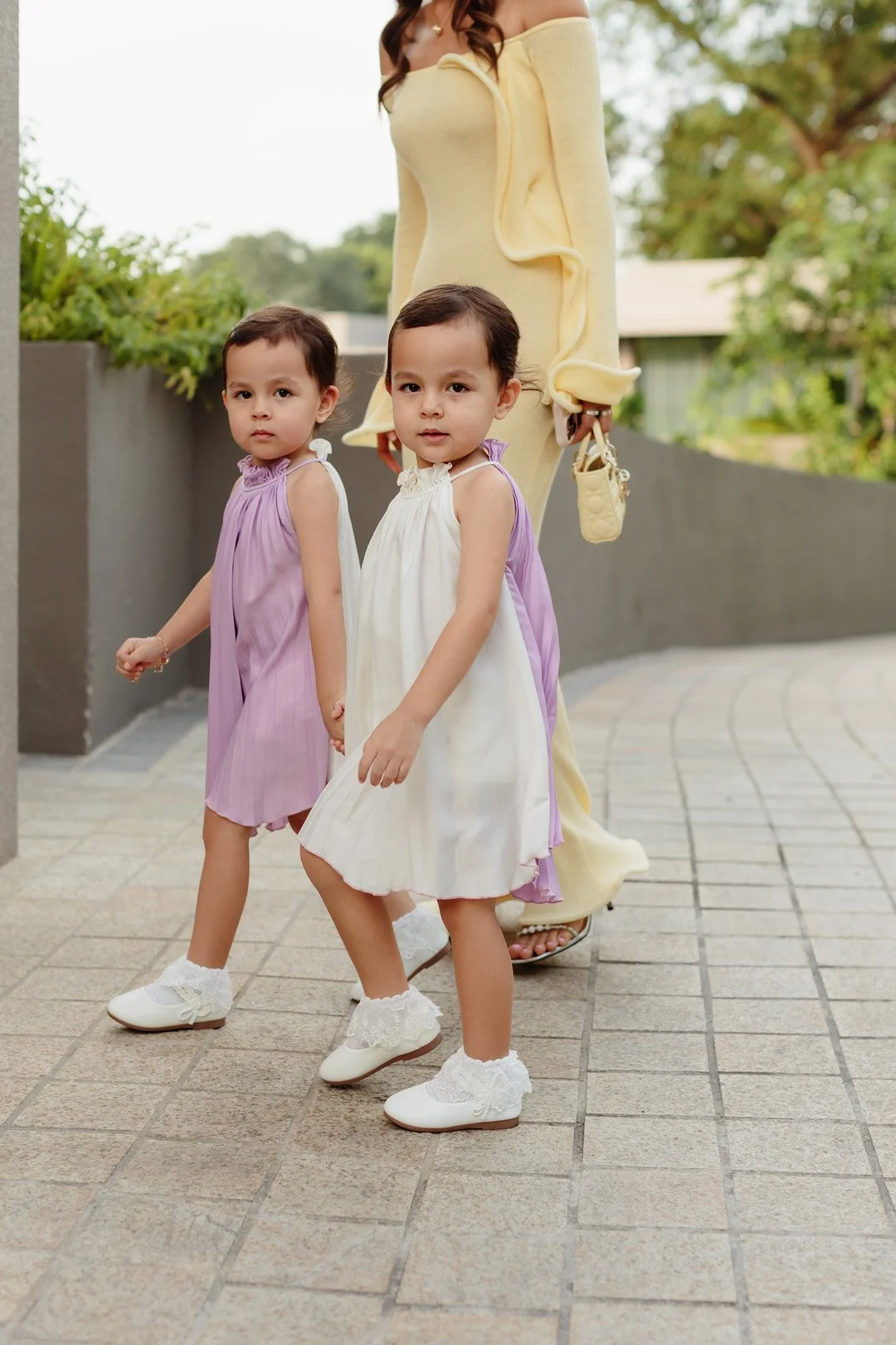 Two young girls holding hands, dressed in pastel-colored dresses, with an adult woman in a yellow dress standing behind them, outdoors on a paved walkway with greenery and trees in the background.