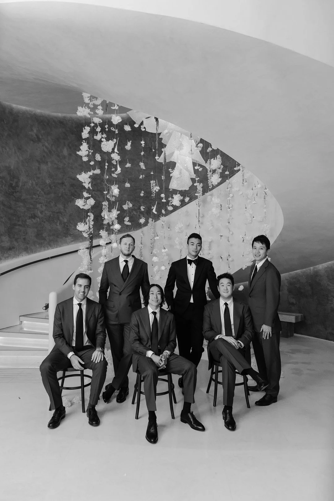 Group of six men in suits and ties, posing indoors against a decorative background with hanging floral arrangements, some sitting on chairs and others standing.