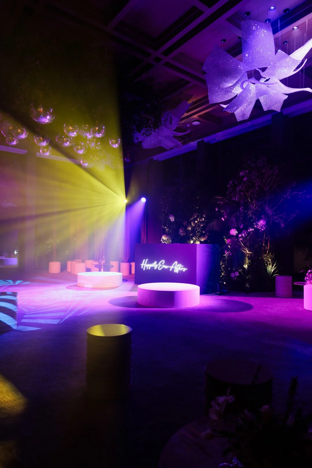 A dimly lit event space with colorful purple, yellow, and pink lighting, decorated with floral arrangements and modern furniture. Neon sign reads "Happily Ever After".