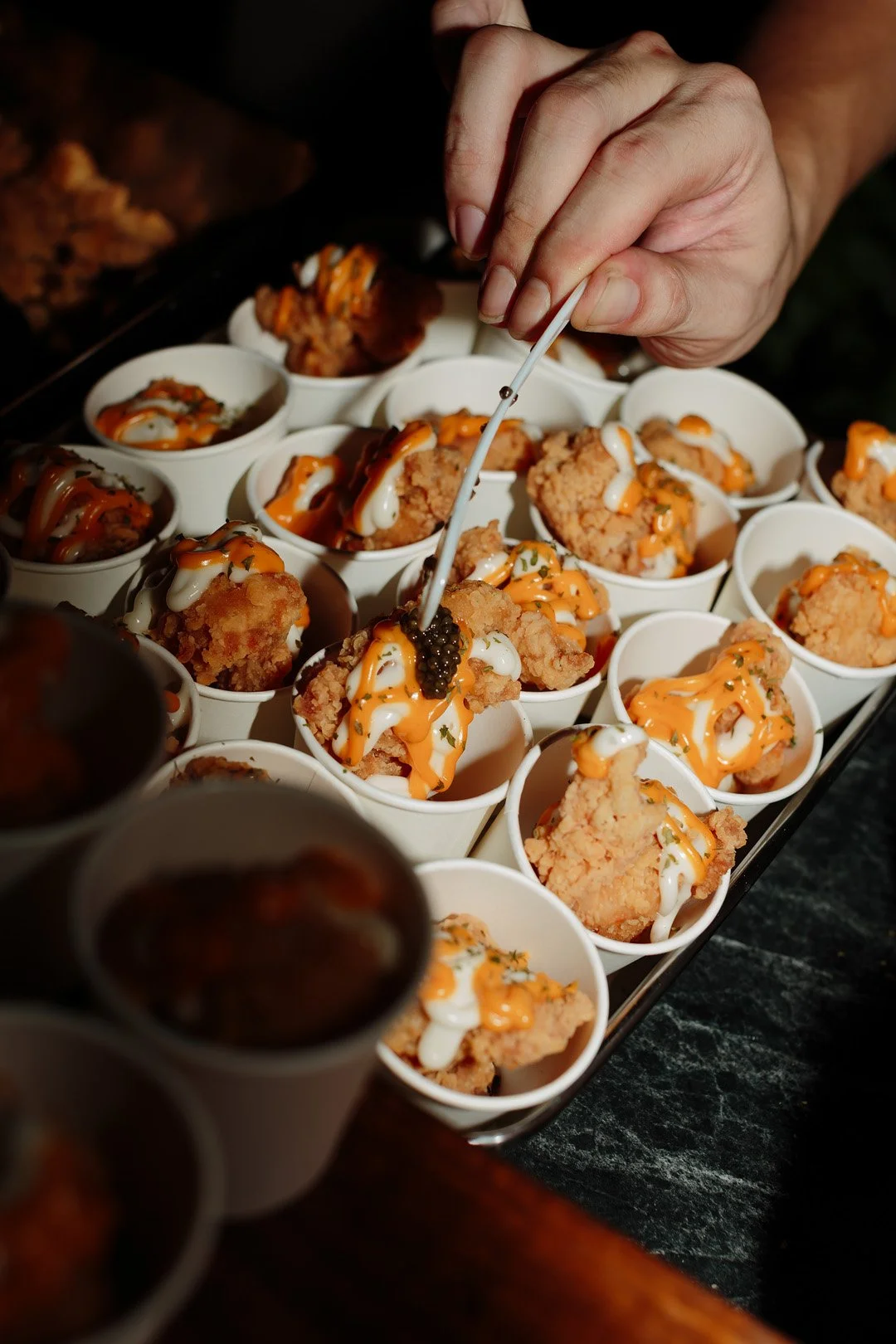 A person using a pair of tongs to place black caviar onto a piece of fried chicken in a white cup, with several similar cups filled with fried chicken, sauces, and garnishes on a serving tray.