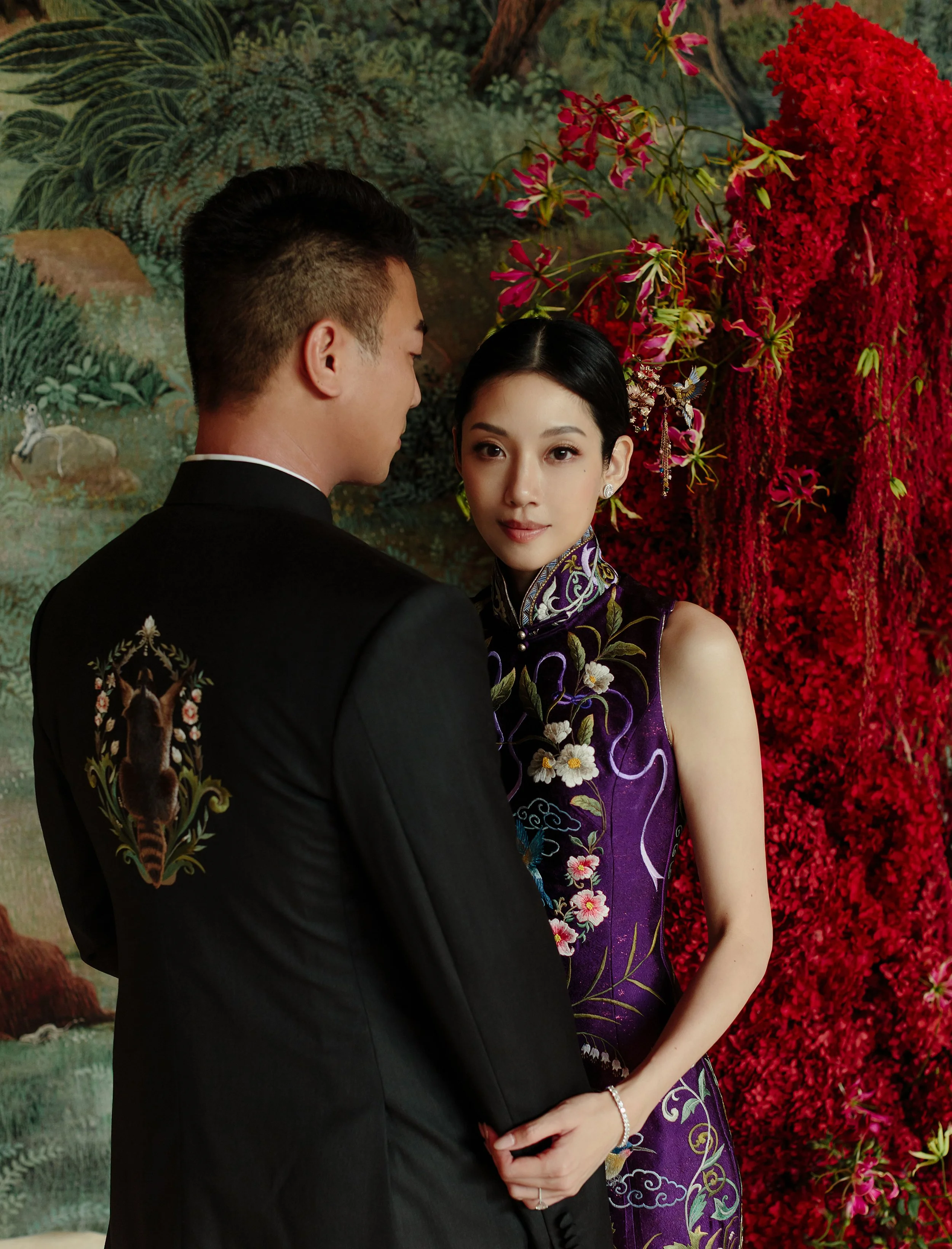 A man and woman in traditional Chinese attire standing close, with the woman looking at the camera and the man facing away, set against a vibrant floral backdrop.