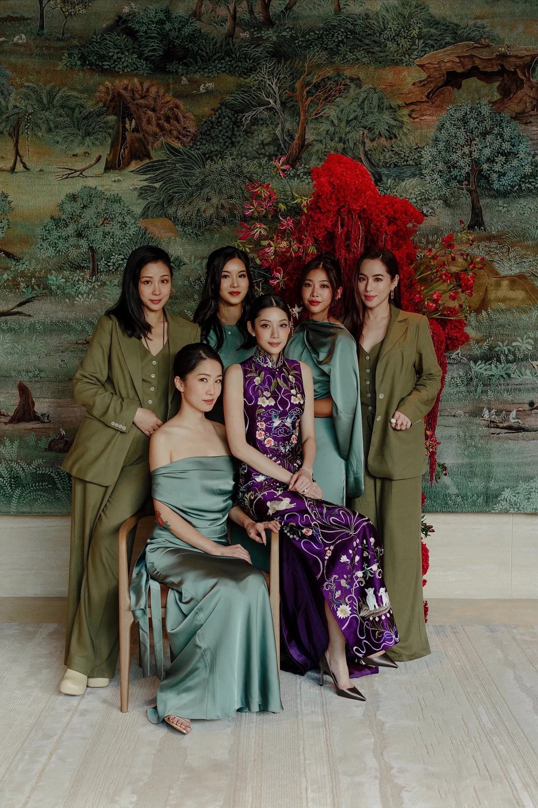Six women posing together in front of a detailed landscape painting with trees, water, and animals, some women are seated while others are standing, dressed in elegant clothing and some in green suits.