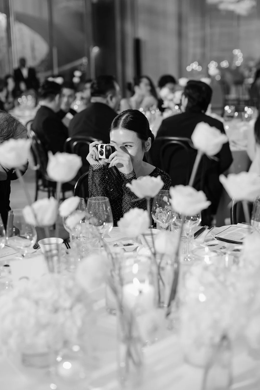 A woman sitting at a banquet table taking a photo with a camera during a formal event, with other guests seated and dining in the background.