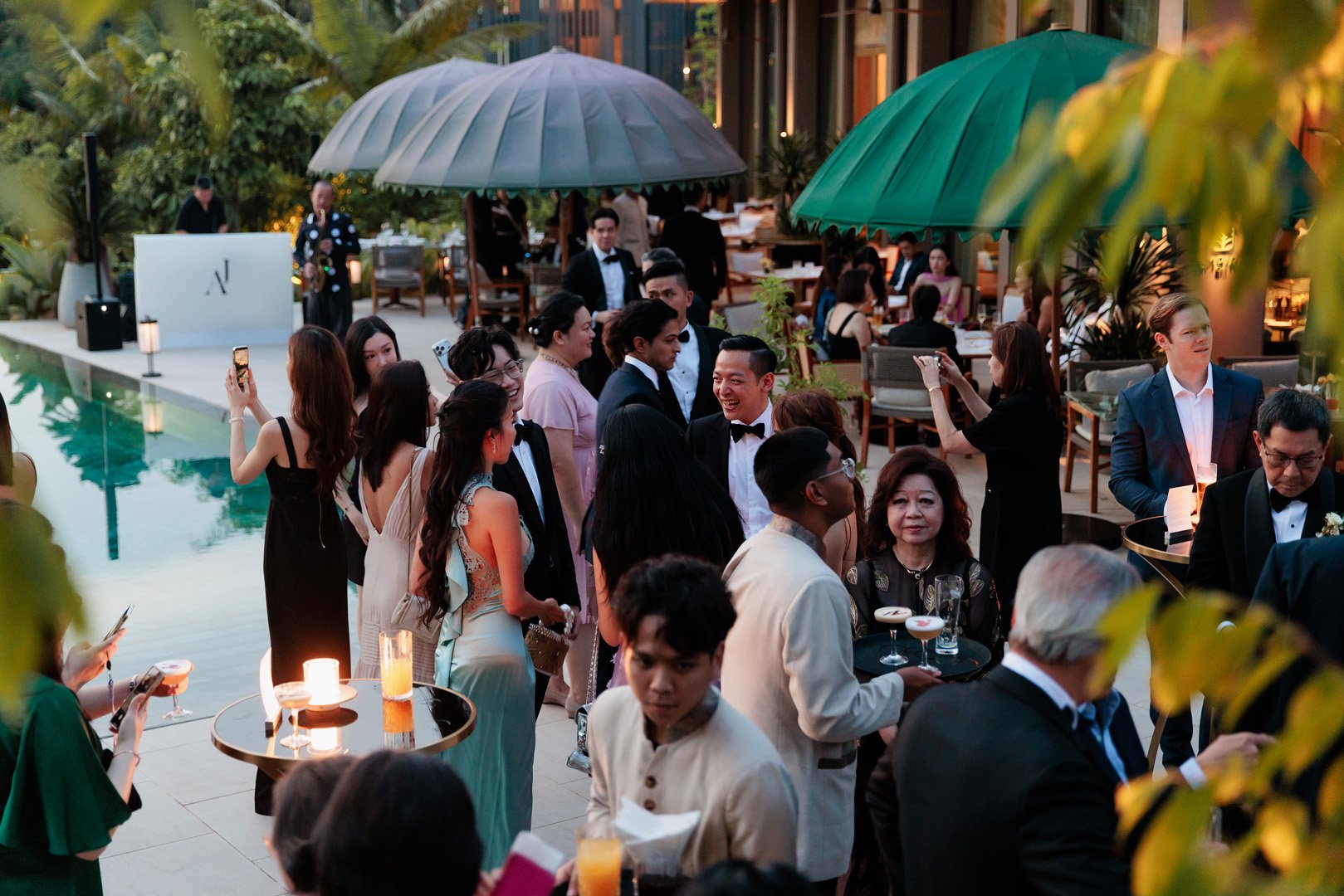 Guests at an outdoor evening event by a pool, dressed in formal attire, with some taking photos and holding drinks under large green umbrellas, with restaurant seating in the background.