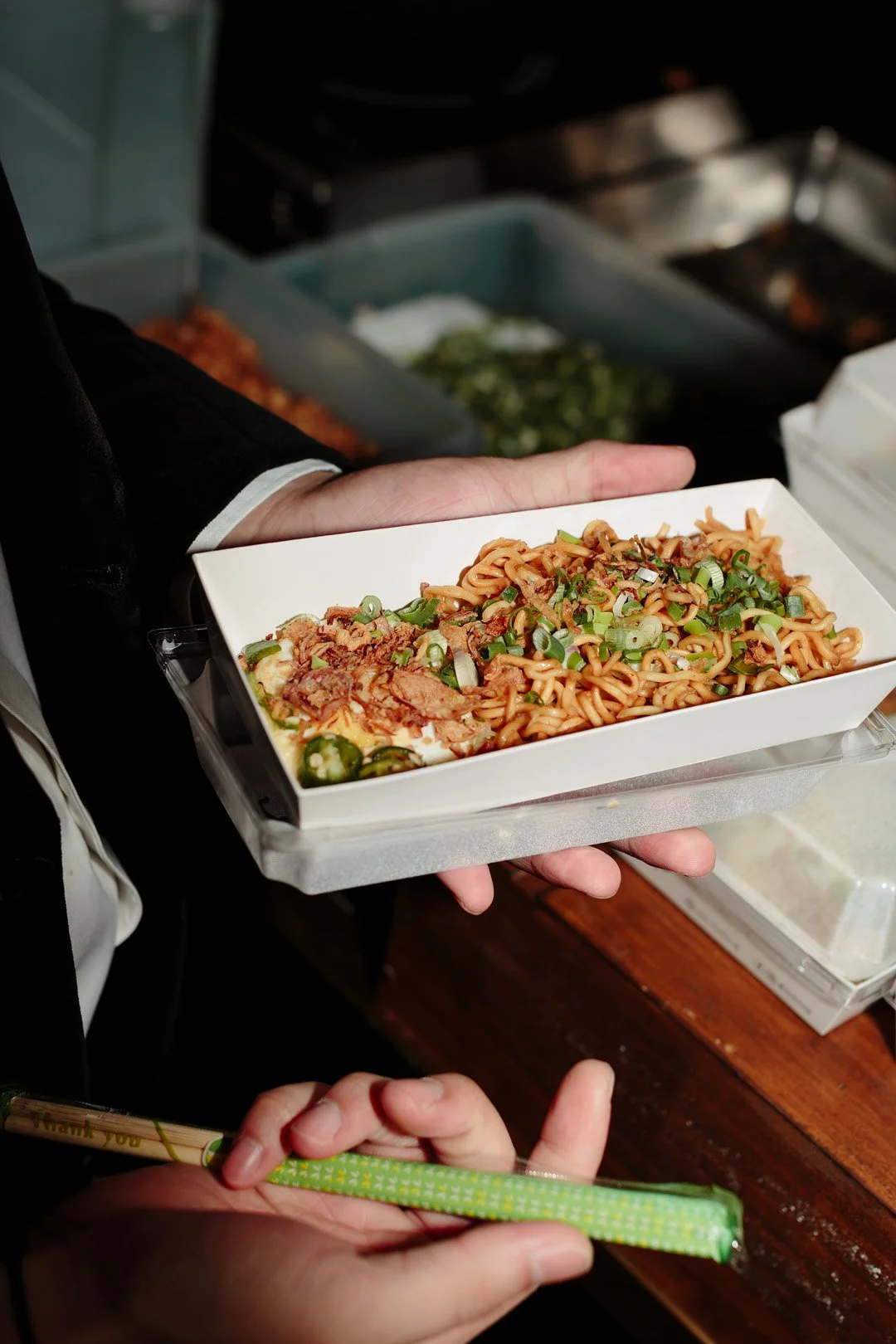 Person holding a white takeout container of Asian stir-fried noodles topped with chopped green onions, with a hand holding a pair of green patterned disposable chopsticks.