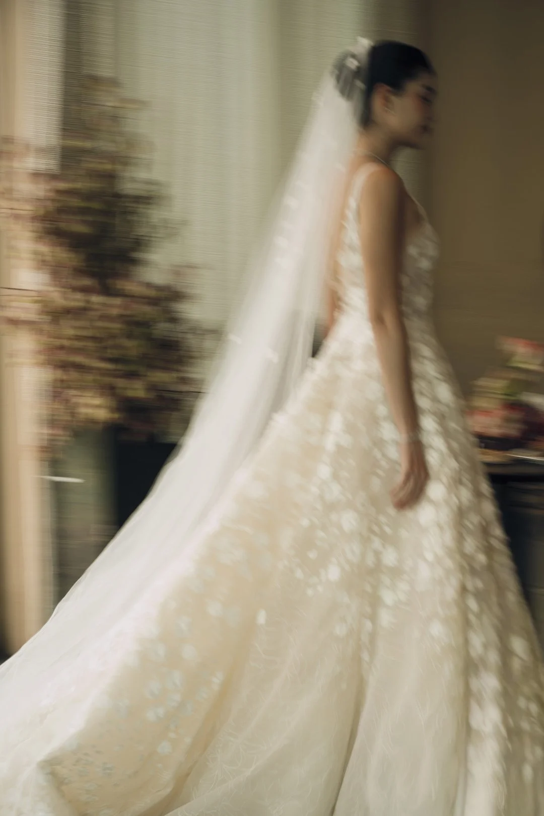A young woman in a wedding dress and veil walking indoors, with a blurred background including flowers and furniture.