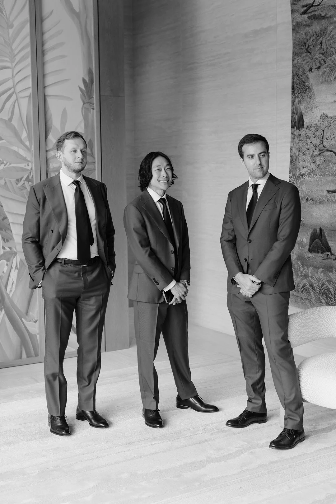 Three business professionals in formal suits standing in a modern indoor setting, two men and one woman, engaged in conversation or waiting.