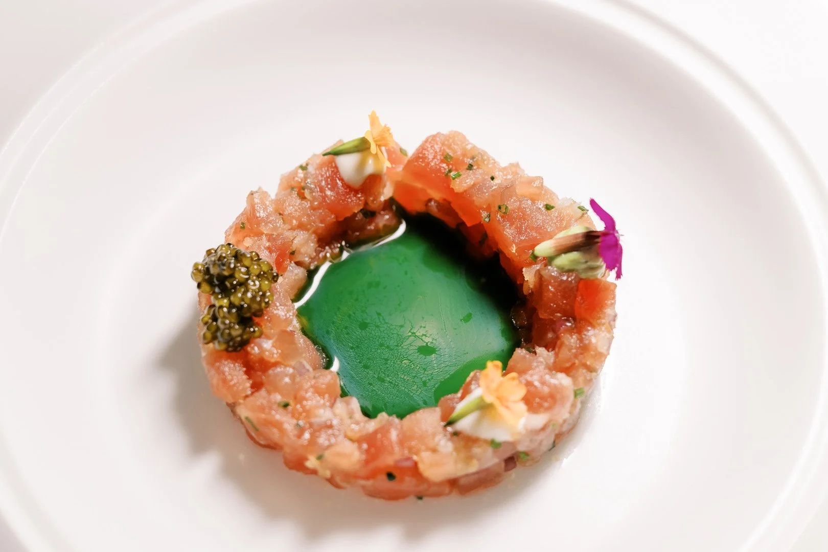 A circular dish of finely chopped raw salmon tartare garnished with edible flowers, green gel in the center, and a small cluster of black caviar on the side, presented on a white plate.