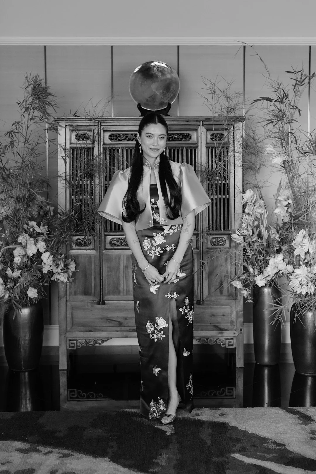 A woman in traditional Asian attire standing in front of a wooden cabinet with floral decorations and potted plants, with a globe above on the wall.
