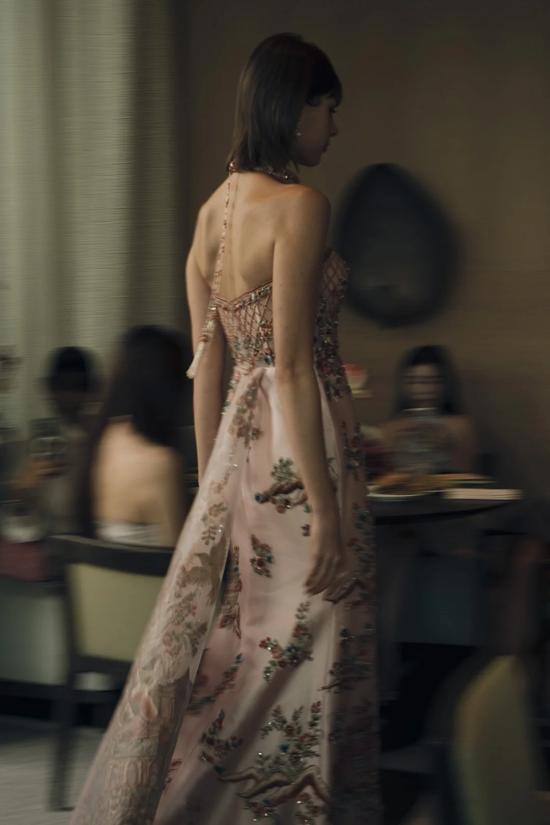 A woman in an ornate, embroidered pink gown with floral patterns walks through a room with other people in the background.