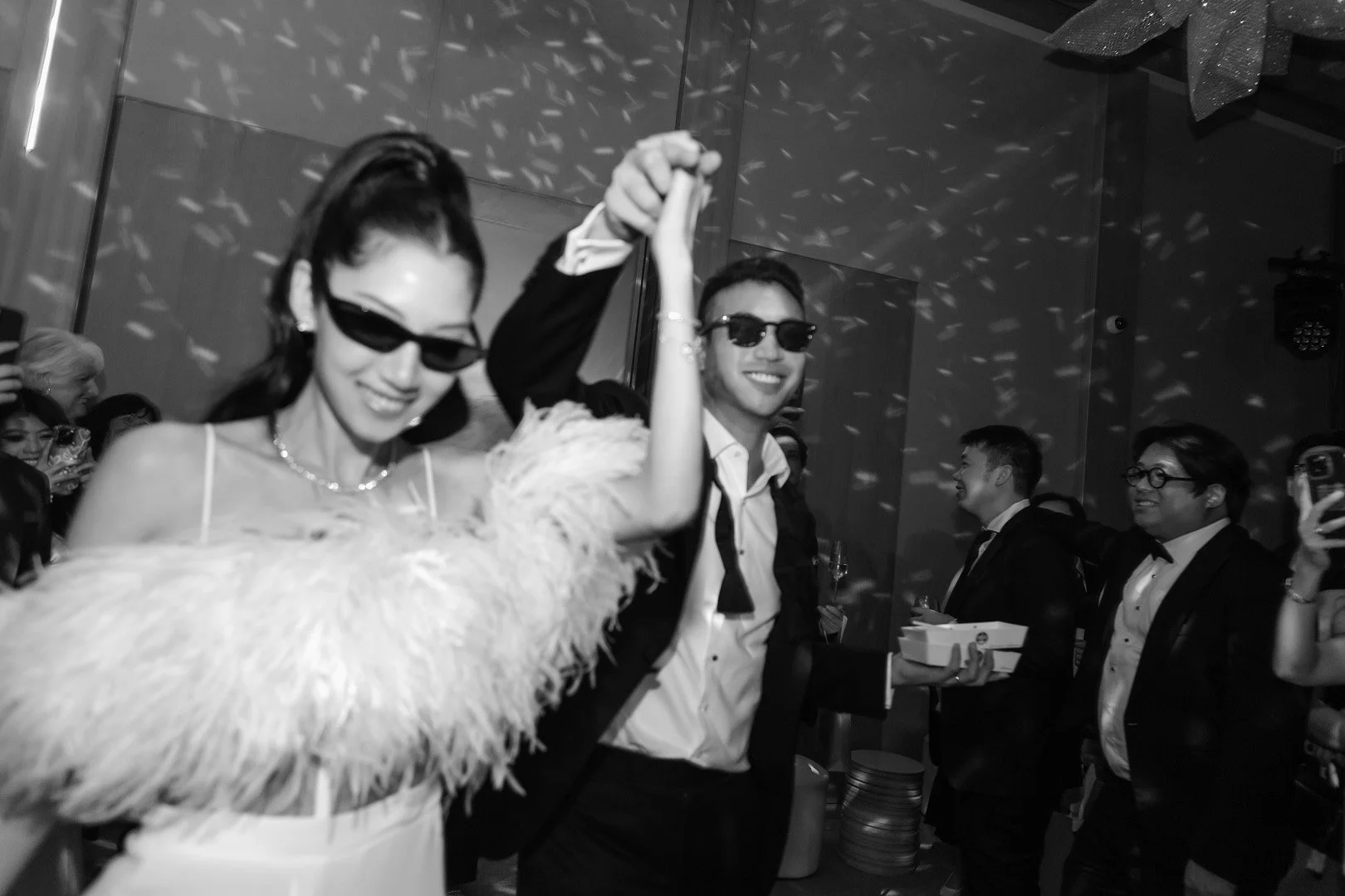 People dancing and celebrating at a party, two individuals in the foreground are holding hands and smiling, wearing sunglasses, surrounded by others in formal attire.