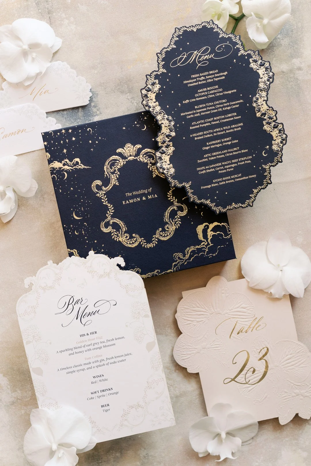 Elegant wedding stationery including a detailed wedding menu, bar menu, and table number card, decorated with white flowers and delicate gold and navy blue accents.