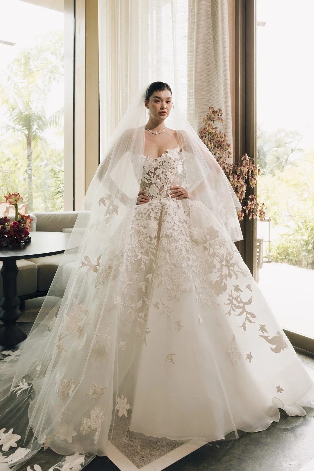 A woman in a wedding dress standing indoors near a large window, with floral decorations and natural light.