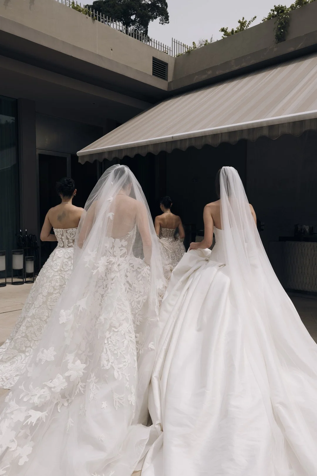 Four women in wedding dresses, viewed from behind, walking outdoors under an overhang with a beige awning.