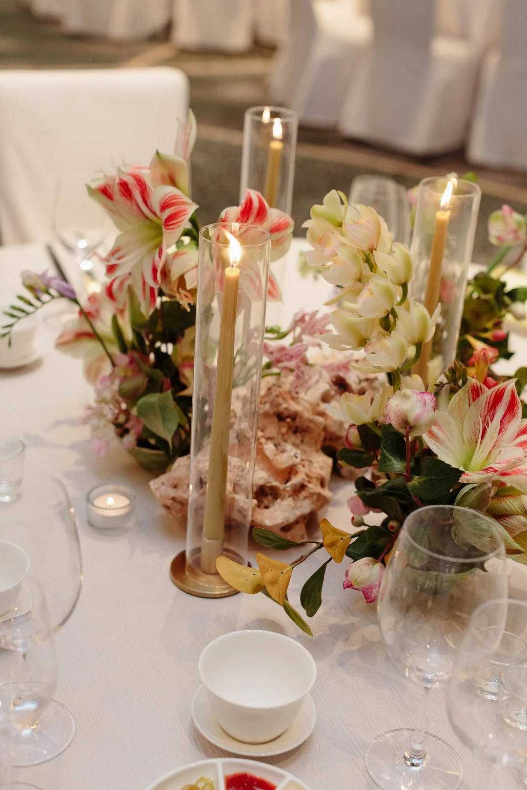 Elegant table centerpiece with white and pink striped lilies, white orchids, and yellow flowers, adorned with tall candles and surrounded by glassware and dishes.
