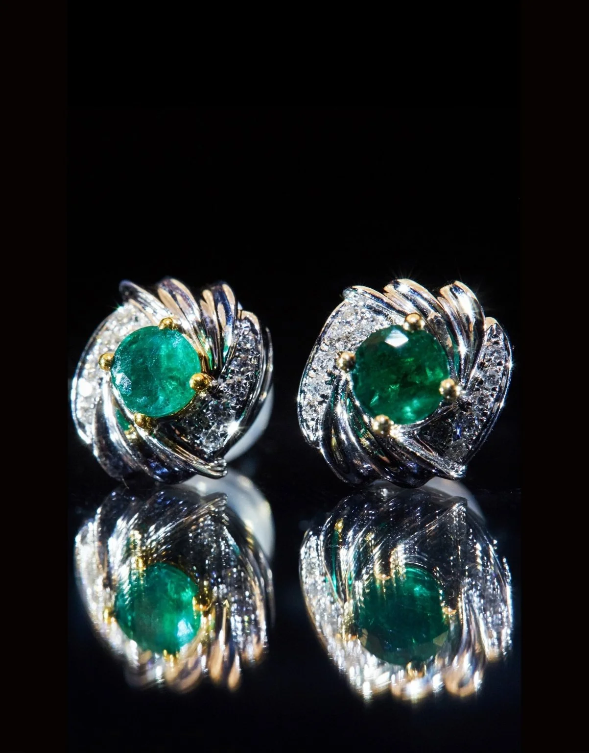 Pair of ornate silver rings with large green gemstones and small diamonds, reflecting on a black surface.