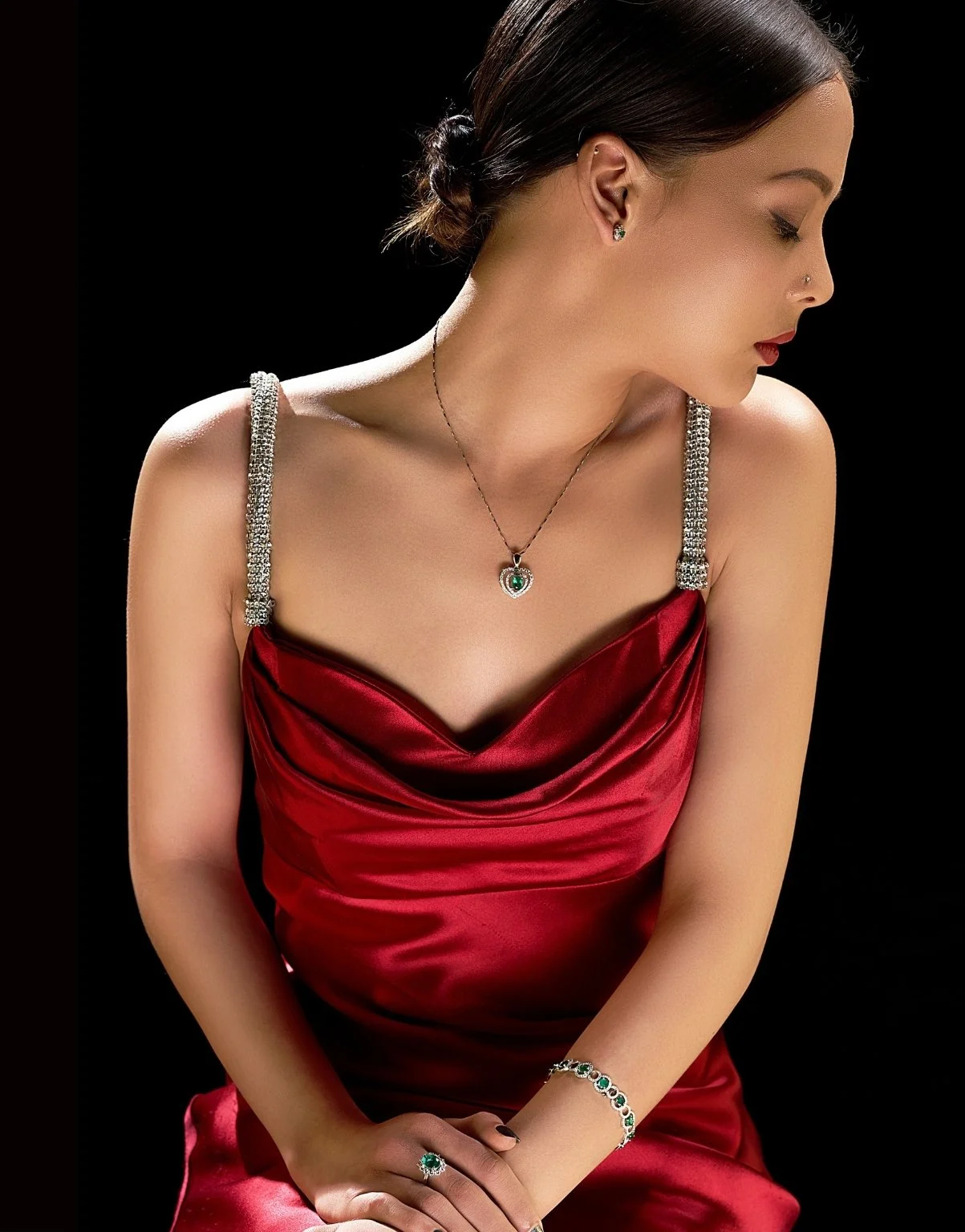 A woman in a red satin dress with silver and green jewelry, including a necklace, bracelet, earrings, and ring, against a black background.