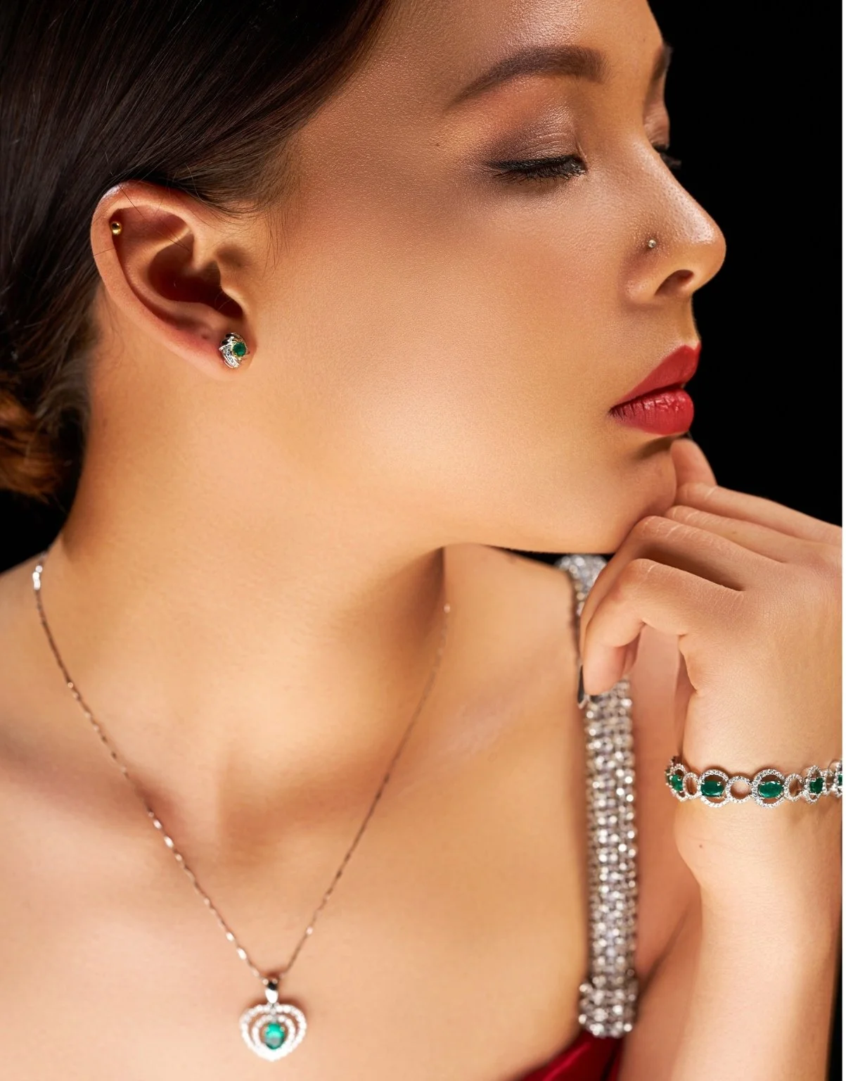 Close-up of a woman with jewelry, including earrings, a necklace, bracelet, and ring, all featuring gemstones, with a black background.