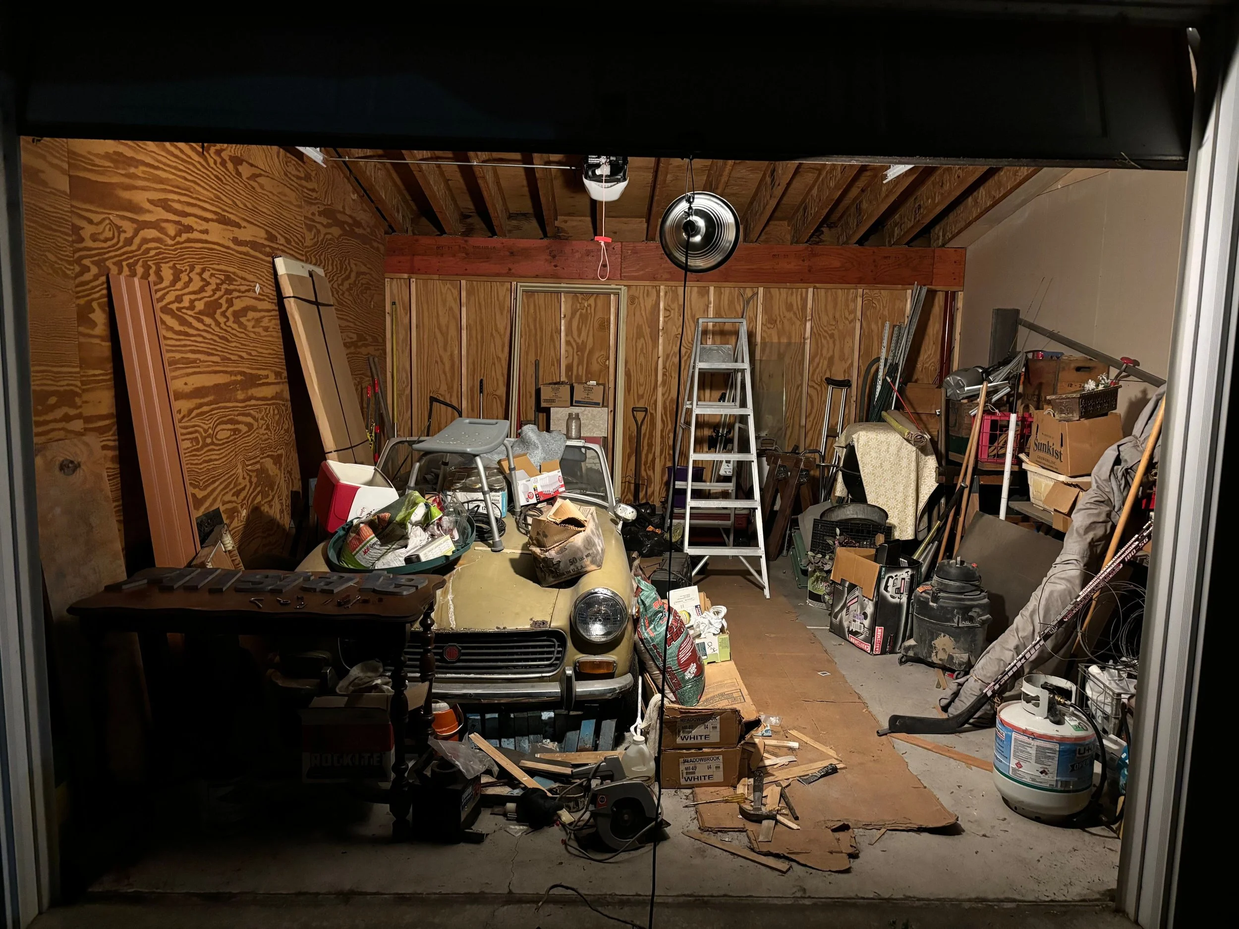 A cluttered garage with various tools, boxes, and an old car in the center, illuminated by a ceiling light.