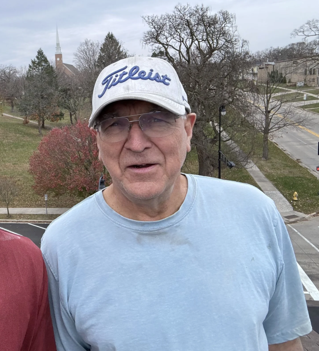A middle-aged man wearing a Titleist cap and glasses, smiling outdoors with trees, a church steeple, and streets in the background.
