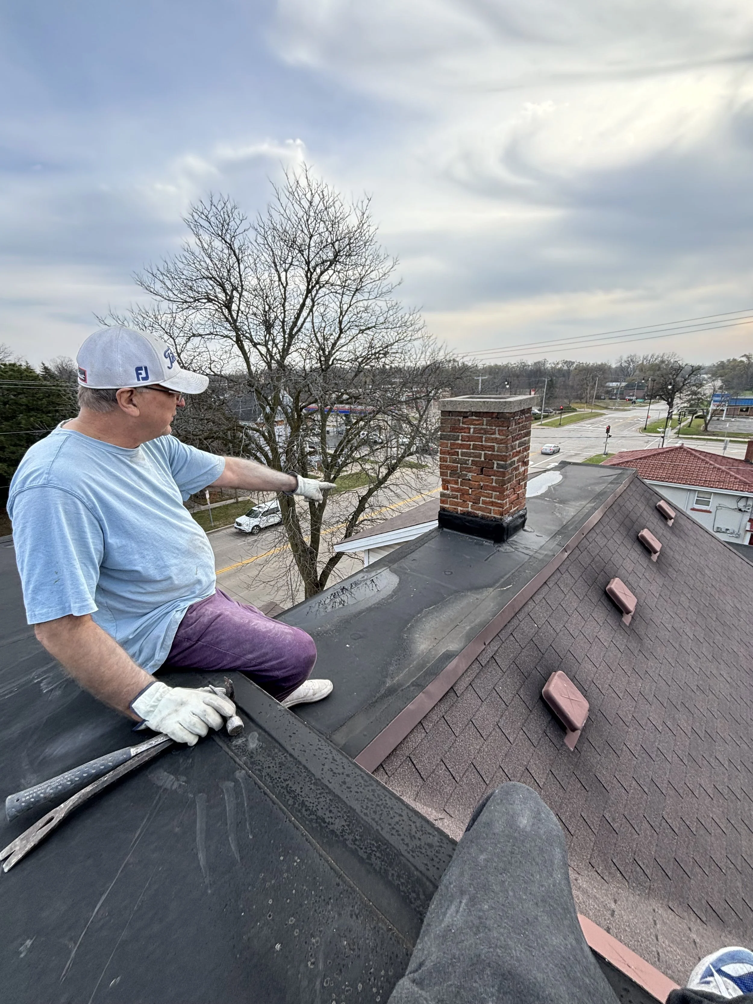 A man working on the roof of a house, sitting on the shingles, wearing gloves, a blue t-shirt, purple pants, and a white cap, with a chimney and cloudy sky in the background.