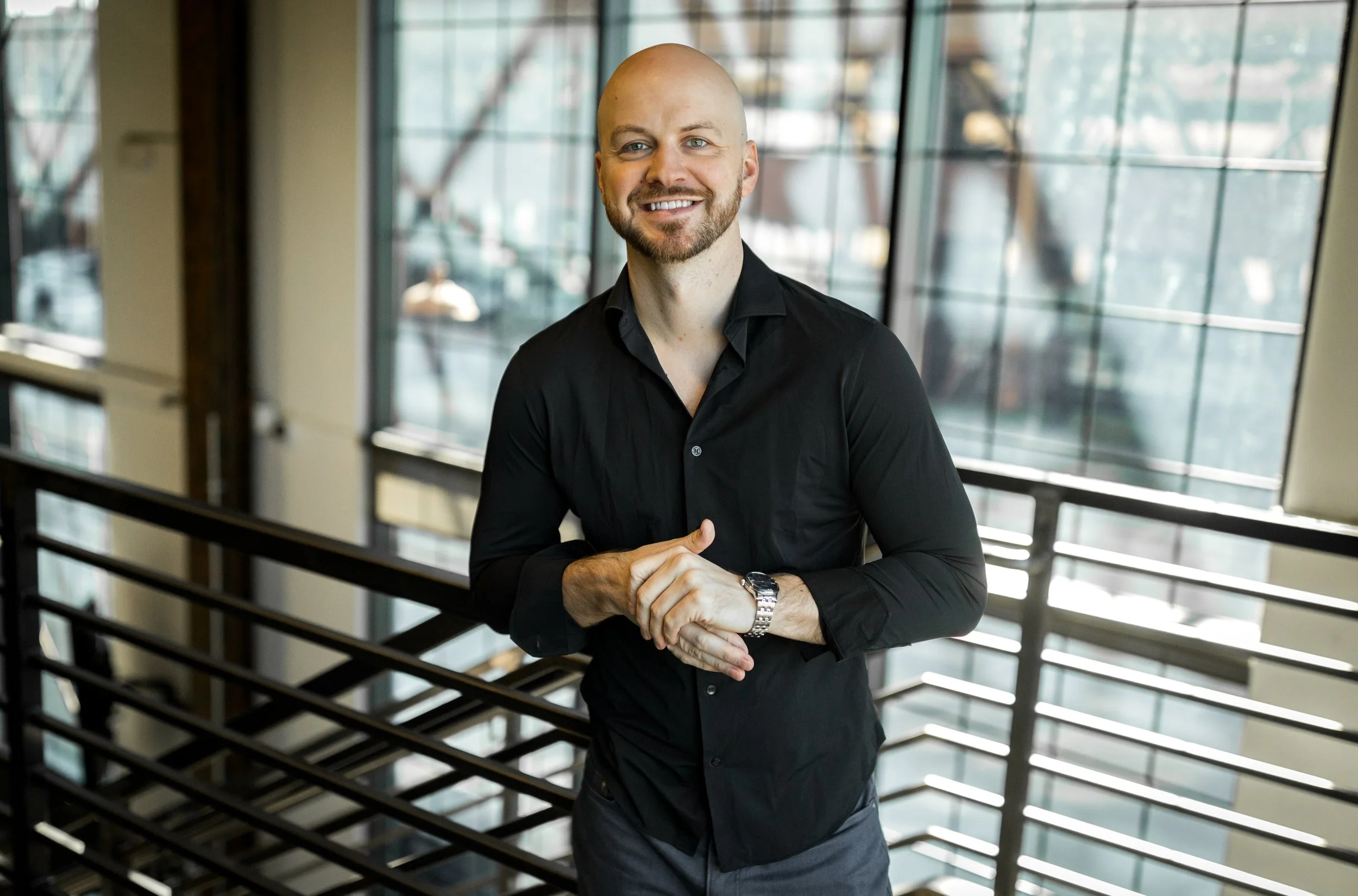 Man with a beard, dressed in a black shirt and gray pants, standing in front of large windows in an office building, smiling and looking at the camera.