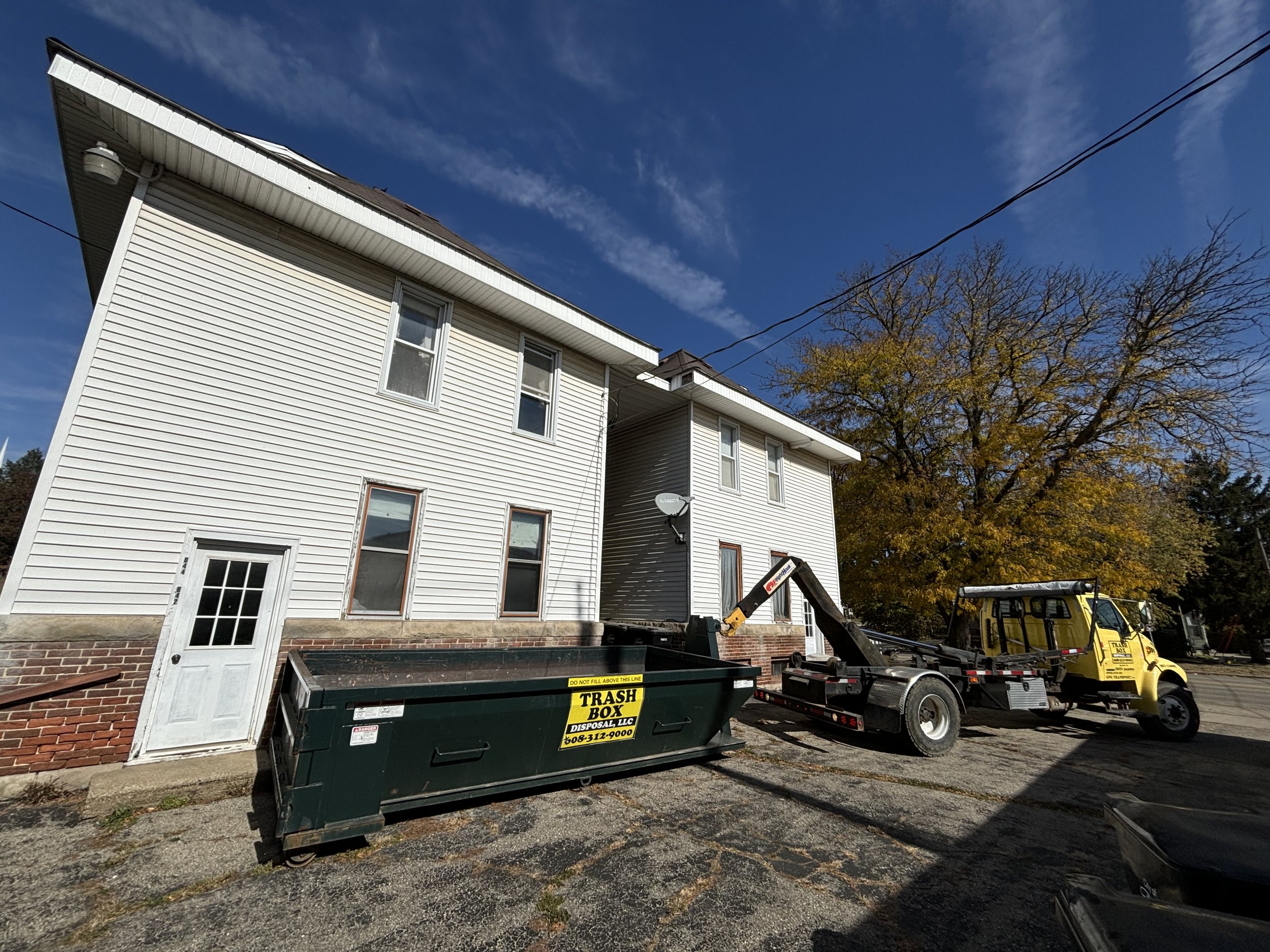 White multi-story house with brick foundation and several windows, a green trash bin labeled 'TRASH BOX,' a tow truck with a yellow cab, and trees with autumn foliage under a partly cloudy sky.