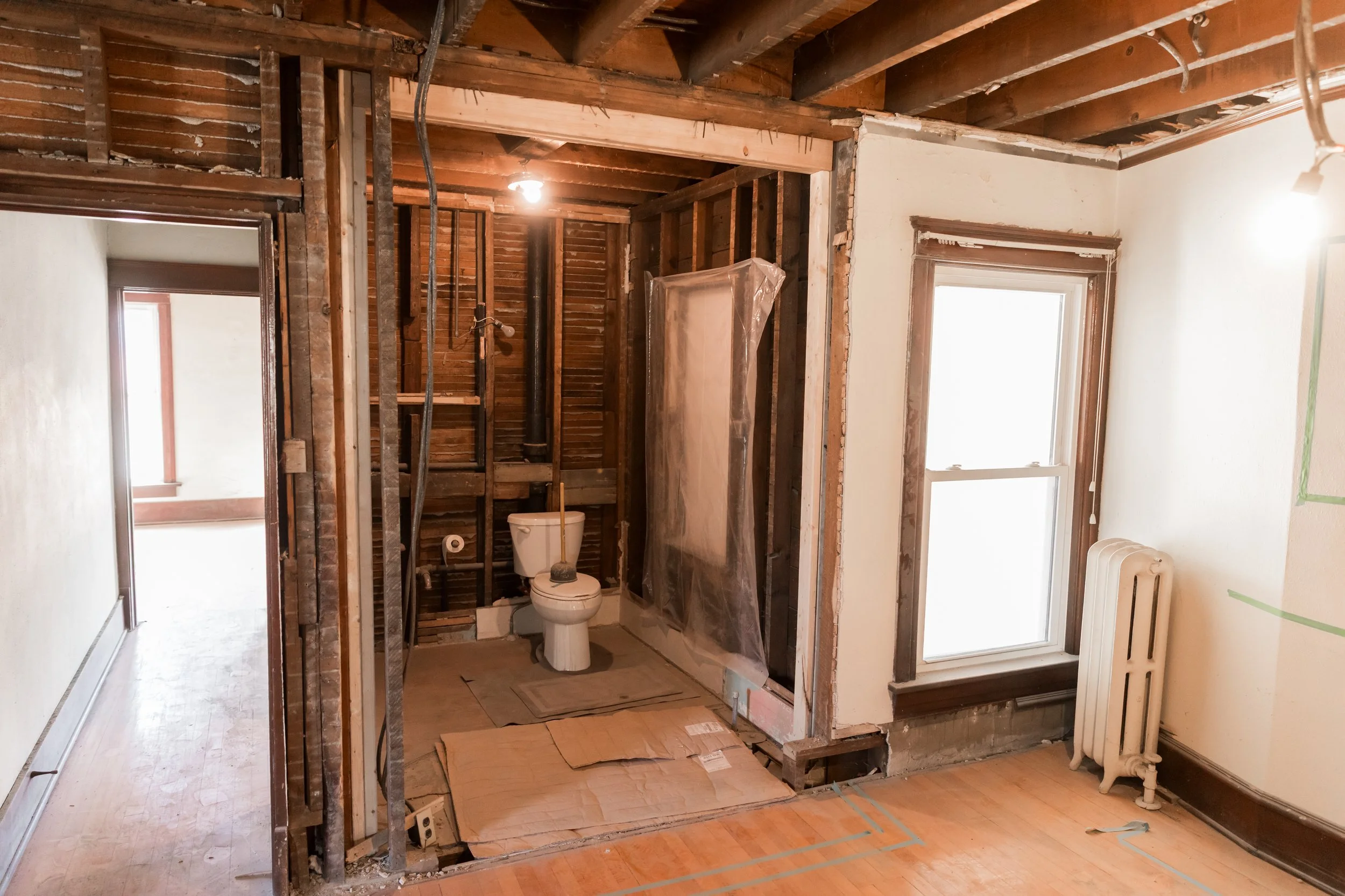 Bathroom under renovation with exposed walls and ceiling, showing a toilet, window, and radiator.