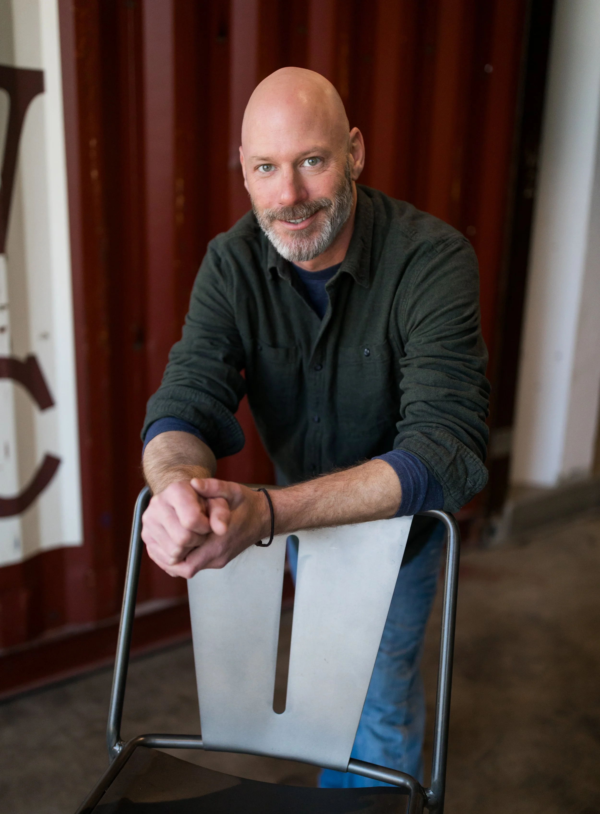 A bald man with a beard leaning on a chair, smiling, indoors with wooden and white walls in the background.