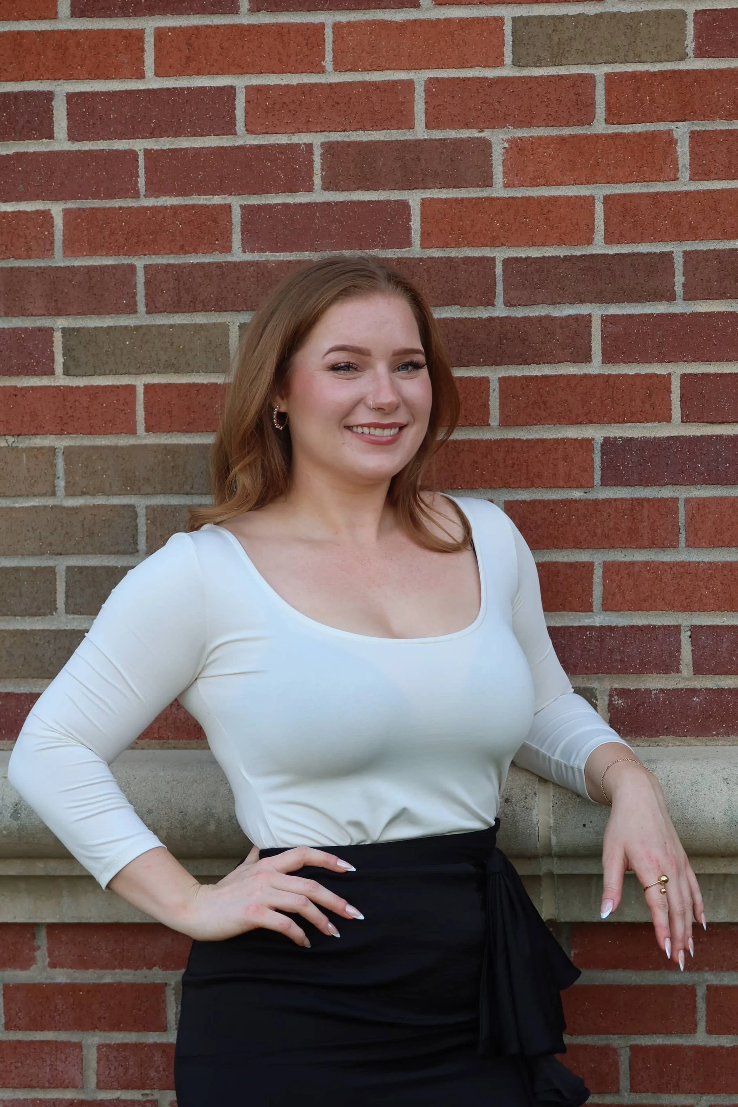 A young woman with auburn hair standing against a brick wall, smiling, wearing a white top and black skirt.