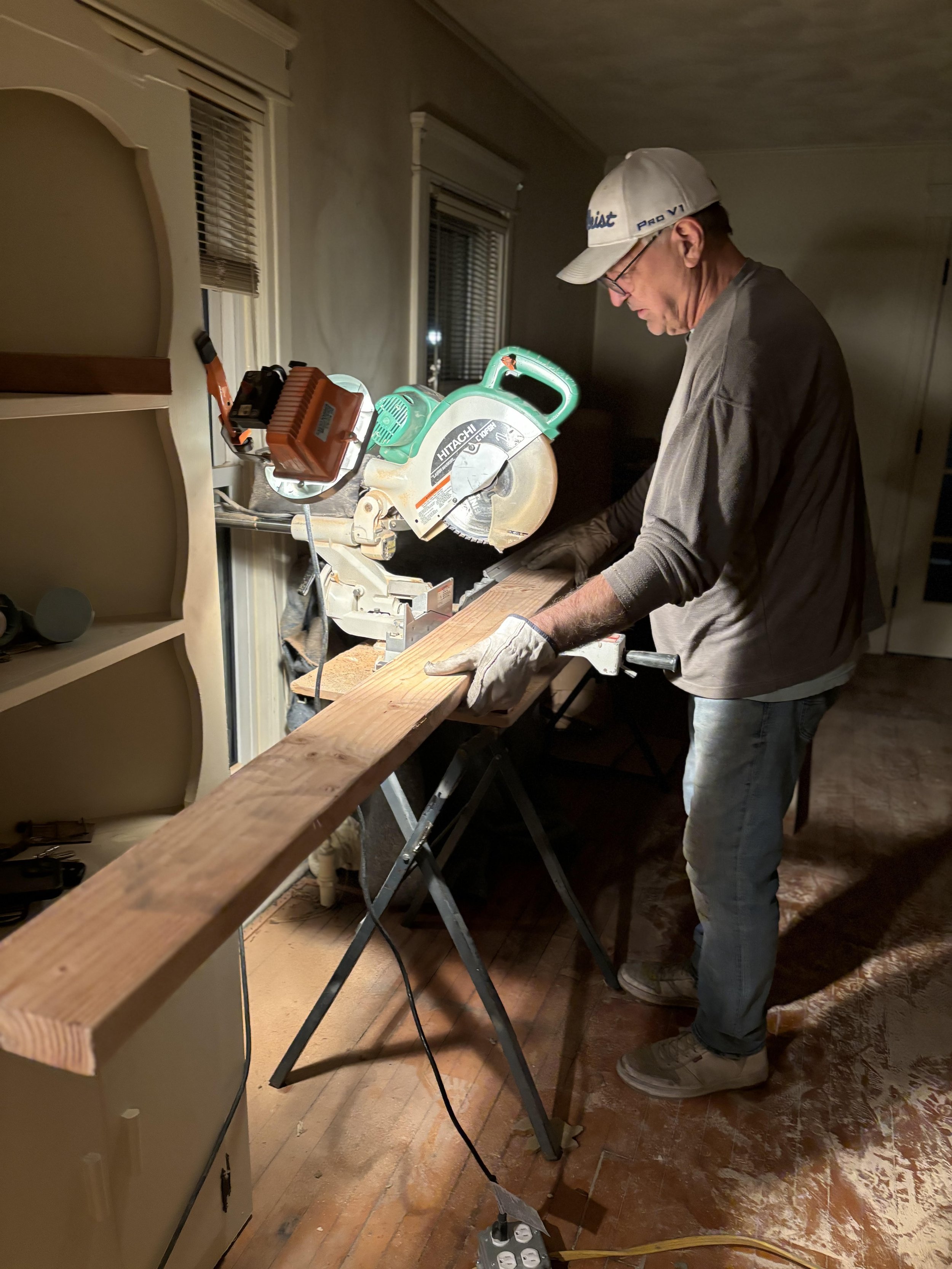 A man wearing a baseball cap, glasses, and work gloves using a circular saw to cut a piece of wood in a dimly lit room.