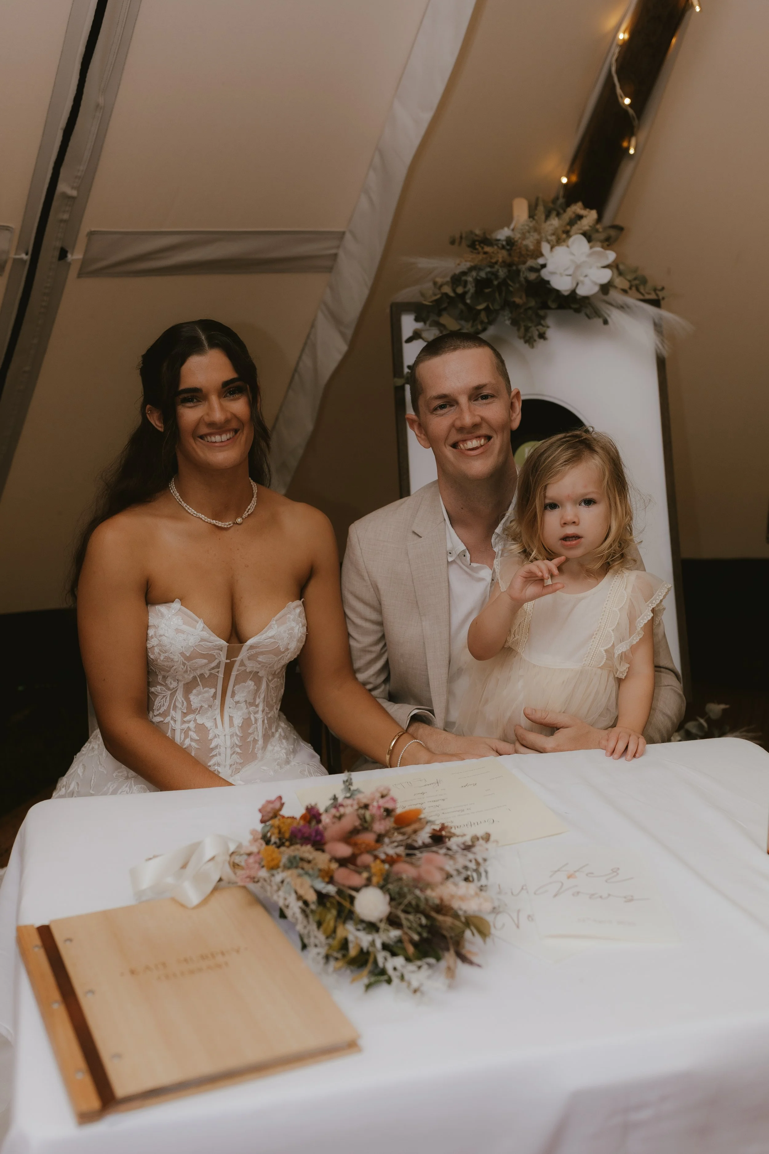 A wedding scene with a bride in a white lace dress, a groom in a beige suit, and a young girl in a cream dress sitting at a table decorated with flowers and signs, inside a tent with string lights and floral arrangements.