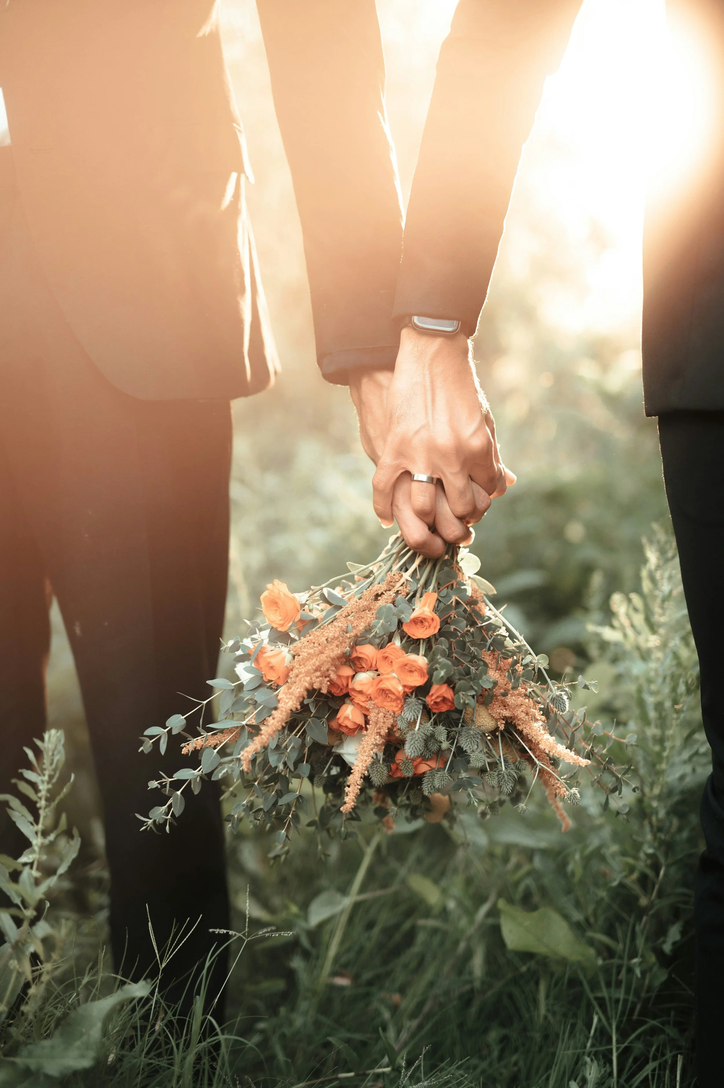 A couple holding hands while standing outdoors, holding a bouquet of orange flowers with greenery, in a sunny, natural setting.