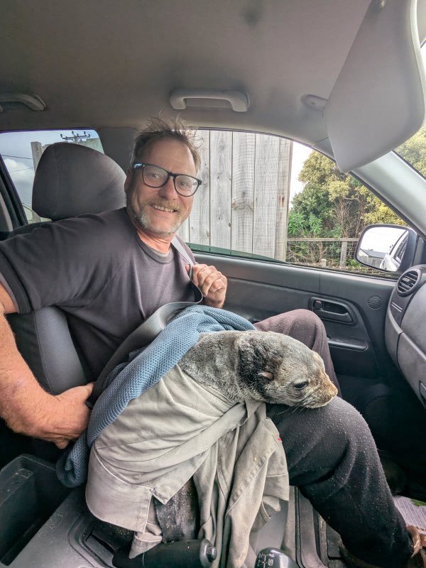 A man wearing glasses, a black helmet, and a black shirt sitting on a red scooter in a parking lot with greenery and a white wooden building in the background.