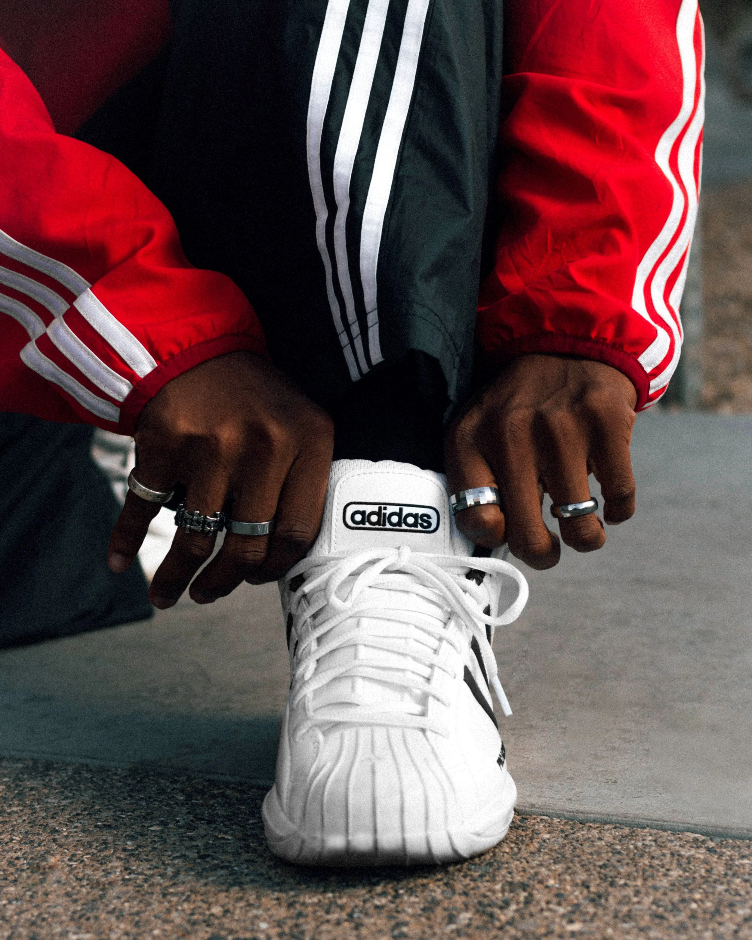 Person wearing a white Adidas sneaker, red and black Adidas jacket, and multiple rings, tying their shoelaces.