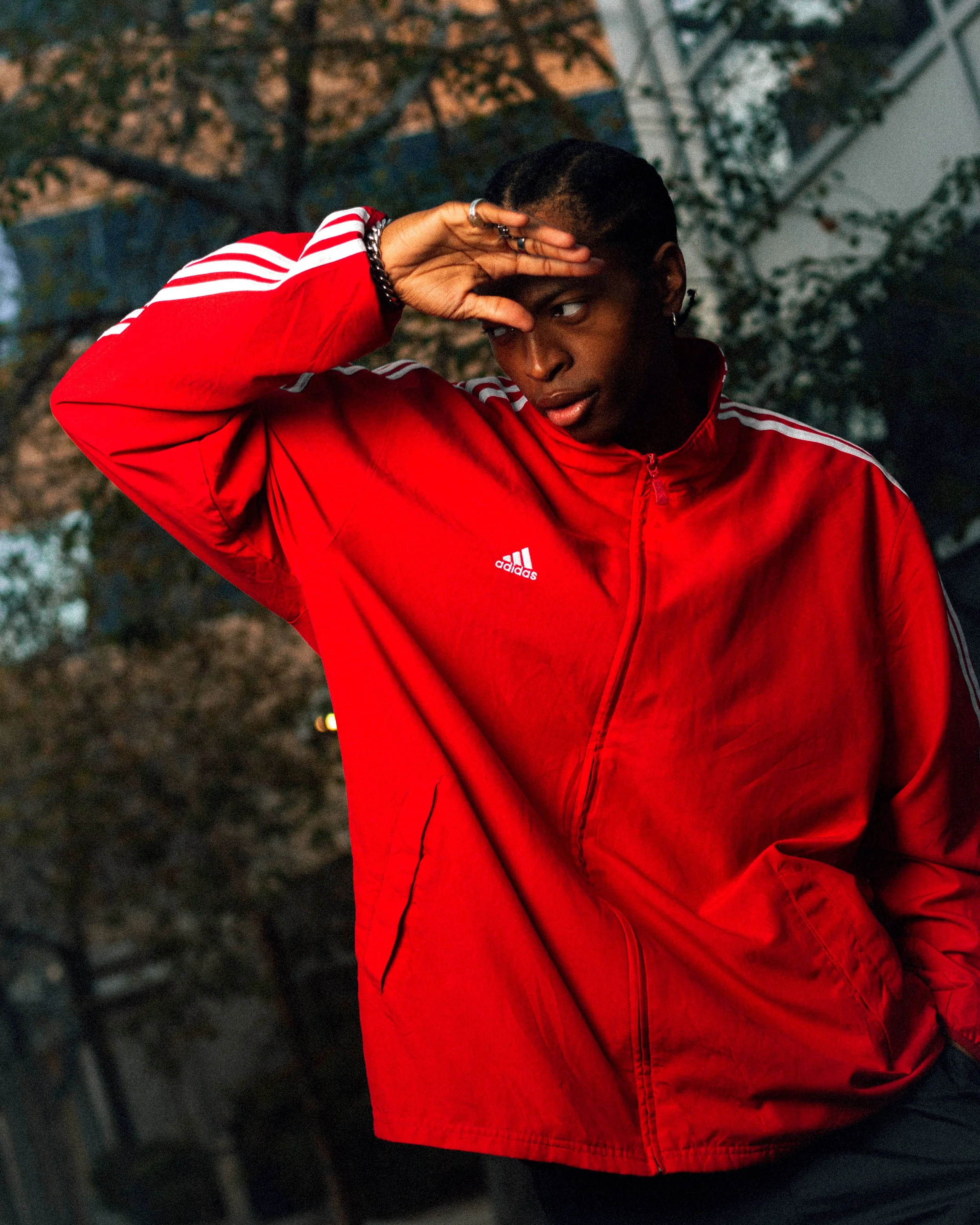 A young man in a red Adidas jacket outside during sunset, shielding his eyes from the light with his hand.