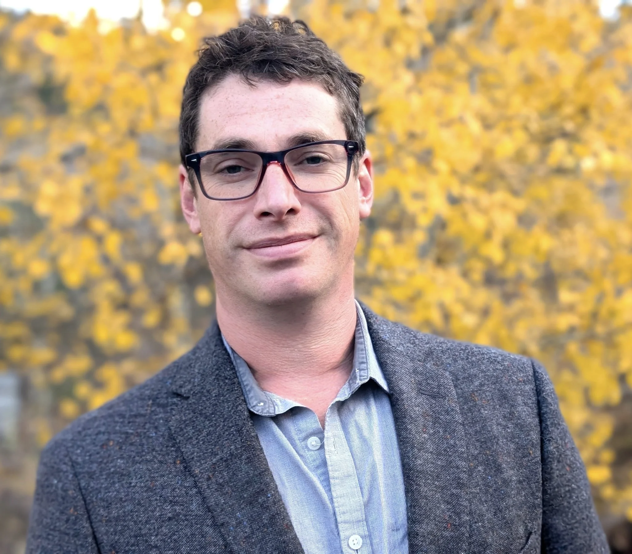 A man with glasses and dark hair, wearing a blazer over a collared shirt, standing outdoors with yellow fall foliage in the background.