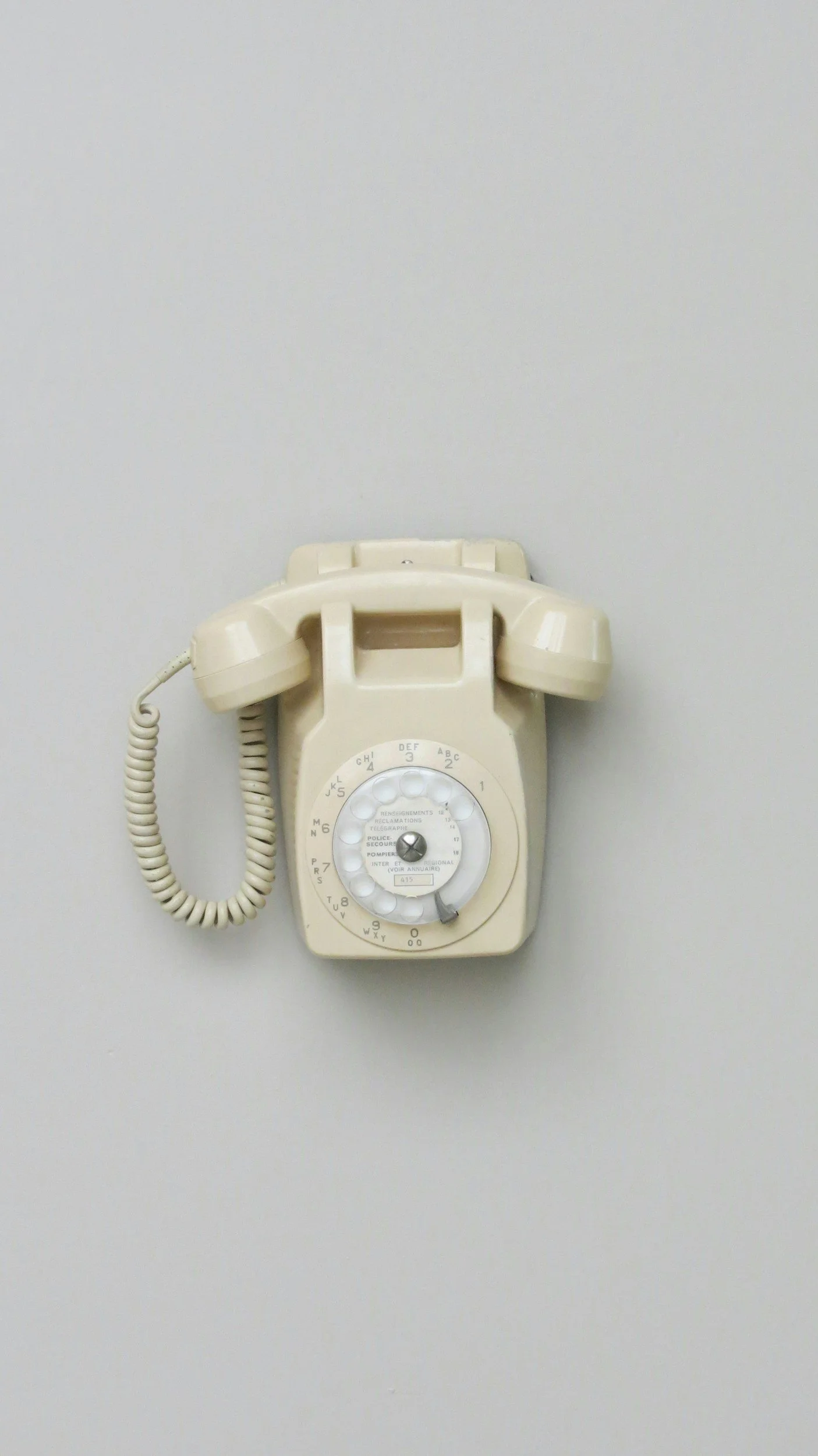 A beige wall-mounted rotary telephone with a coiled cord.