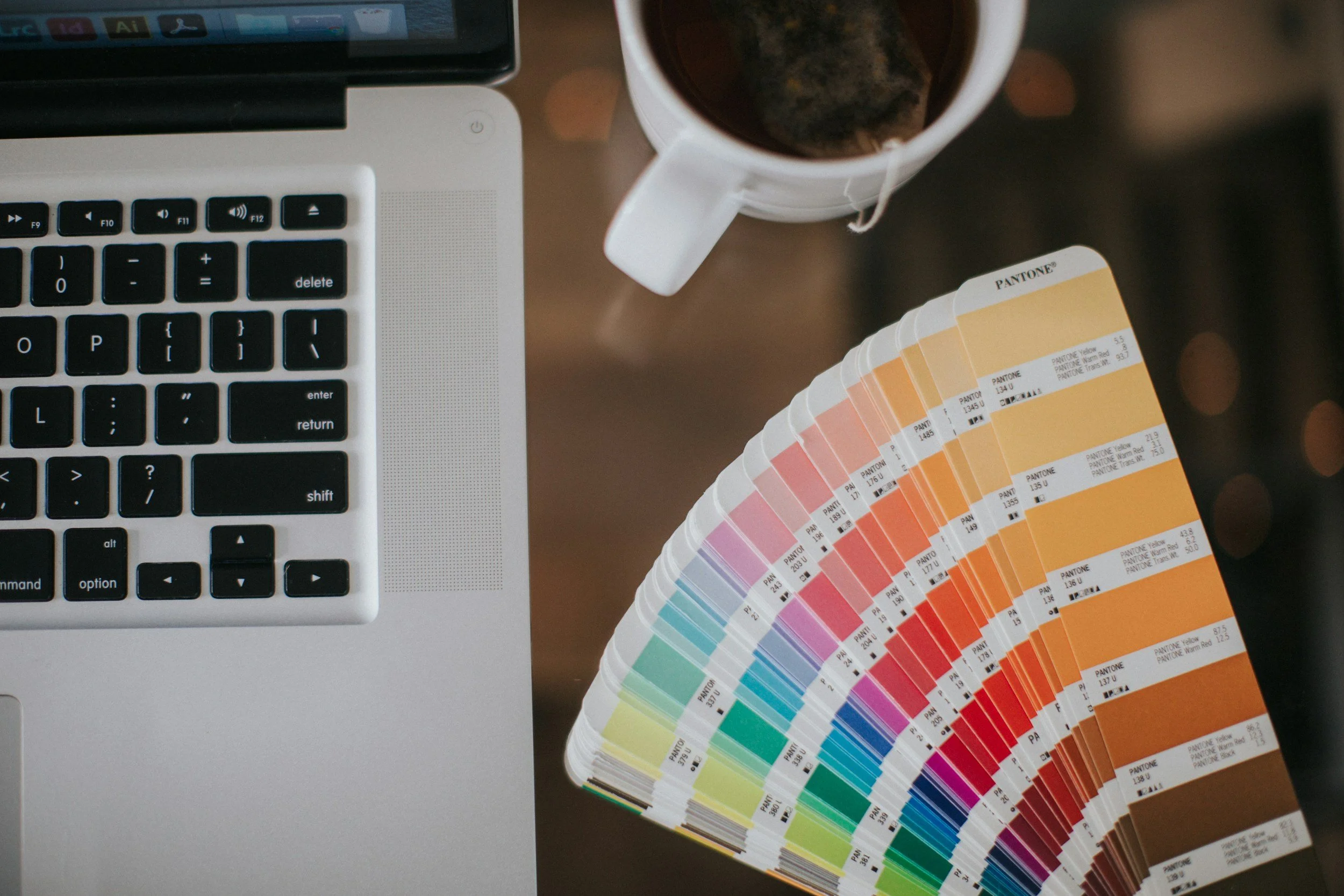 A MacBook laptop with a silver keyboard next to a color palette fan deck and a cup of tea or coffee on a wooden table.