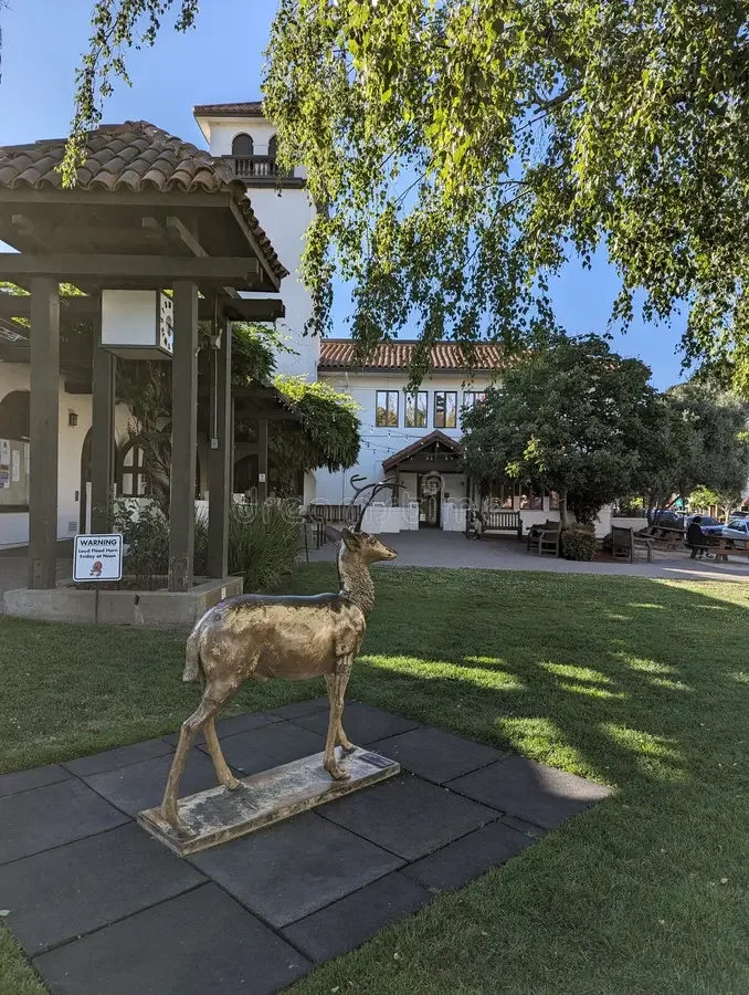 A life-sized deer statue on a paved walkway in a park, with a building and trees in the background, under a blue sky.