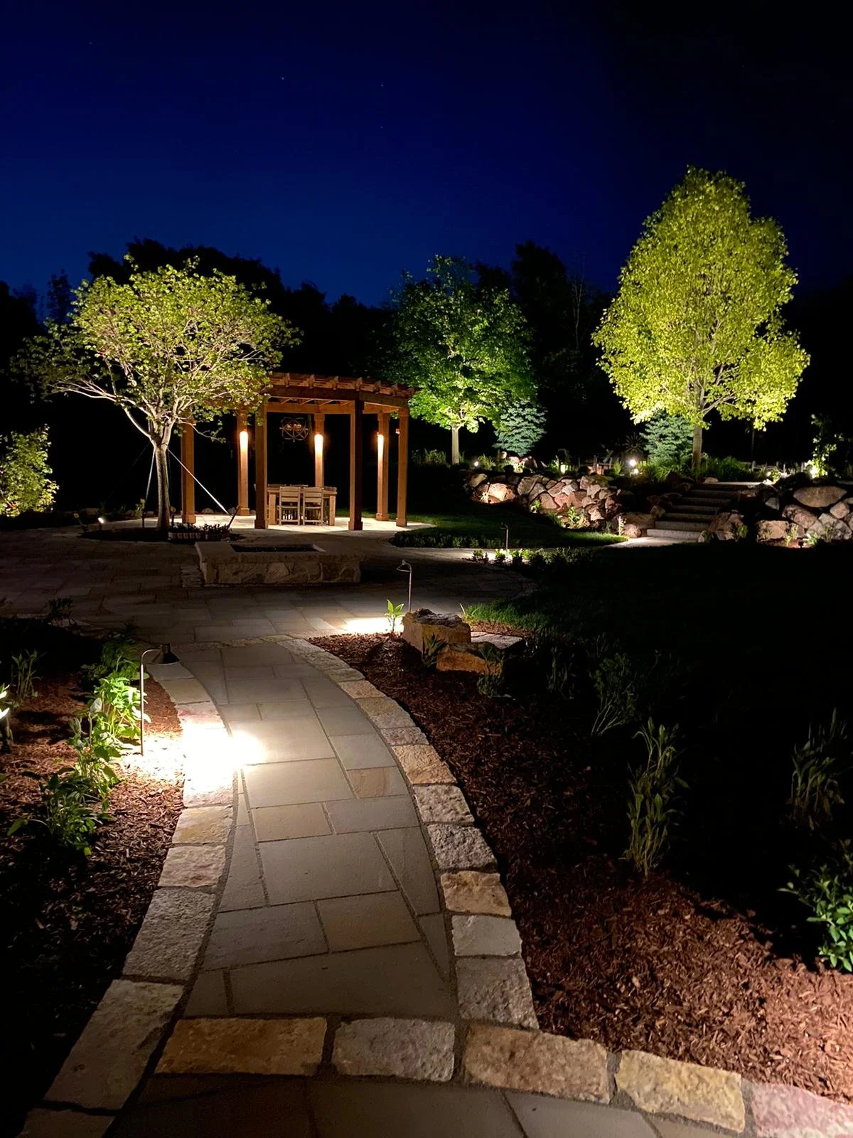 Nighttime view of a landscaped backyard with illuminated trees, a stone pathway, a wooden pergola with chairs, and garden lights.