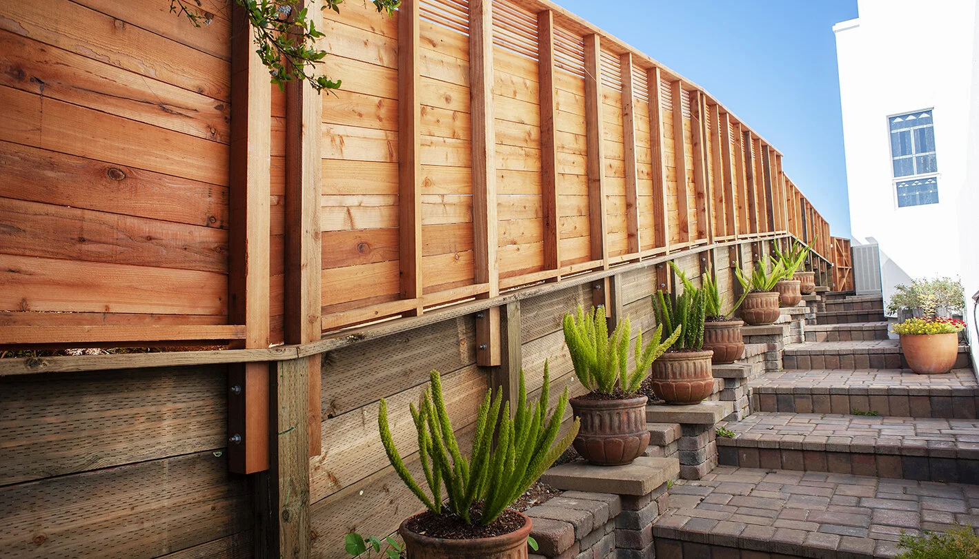 A backyard at sunset with a tall wooden privacy fence, tiered brick steps, and potted plants including cacti and succulents along the stairs.