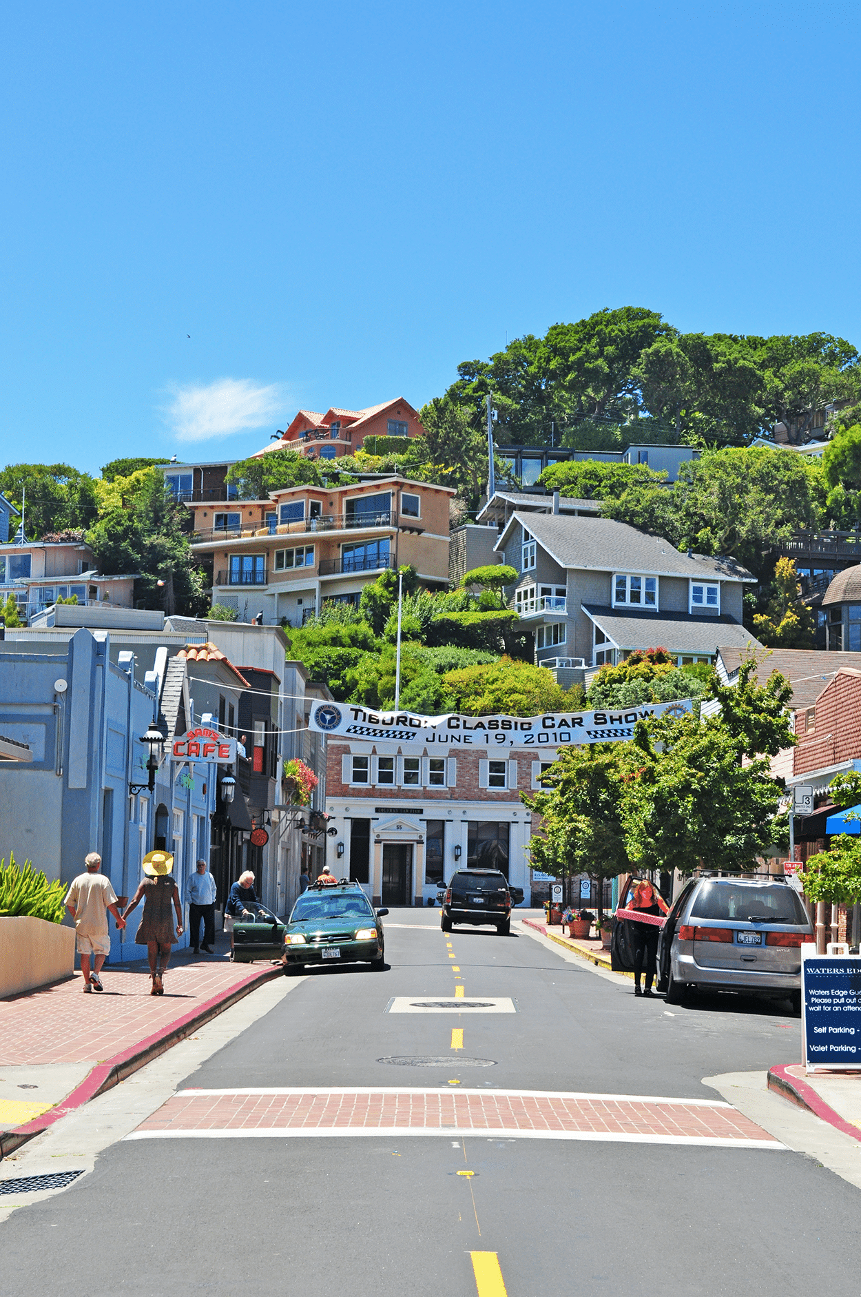 Street scene in a hilly area with colorful buildings, people walking, and cars parked. A banner reads 'Tiburon Classic Car Show June 19, 2010'.