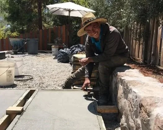 Person wearing a straw hat, smiling, working on a concrete pathway outdoors