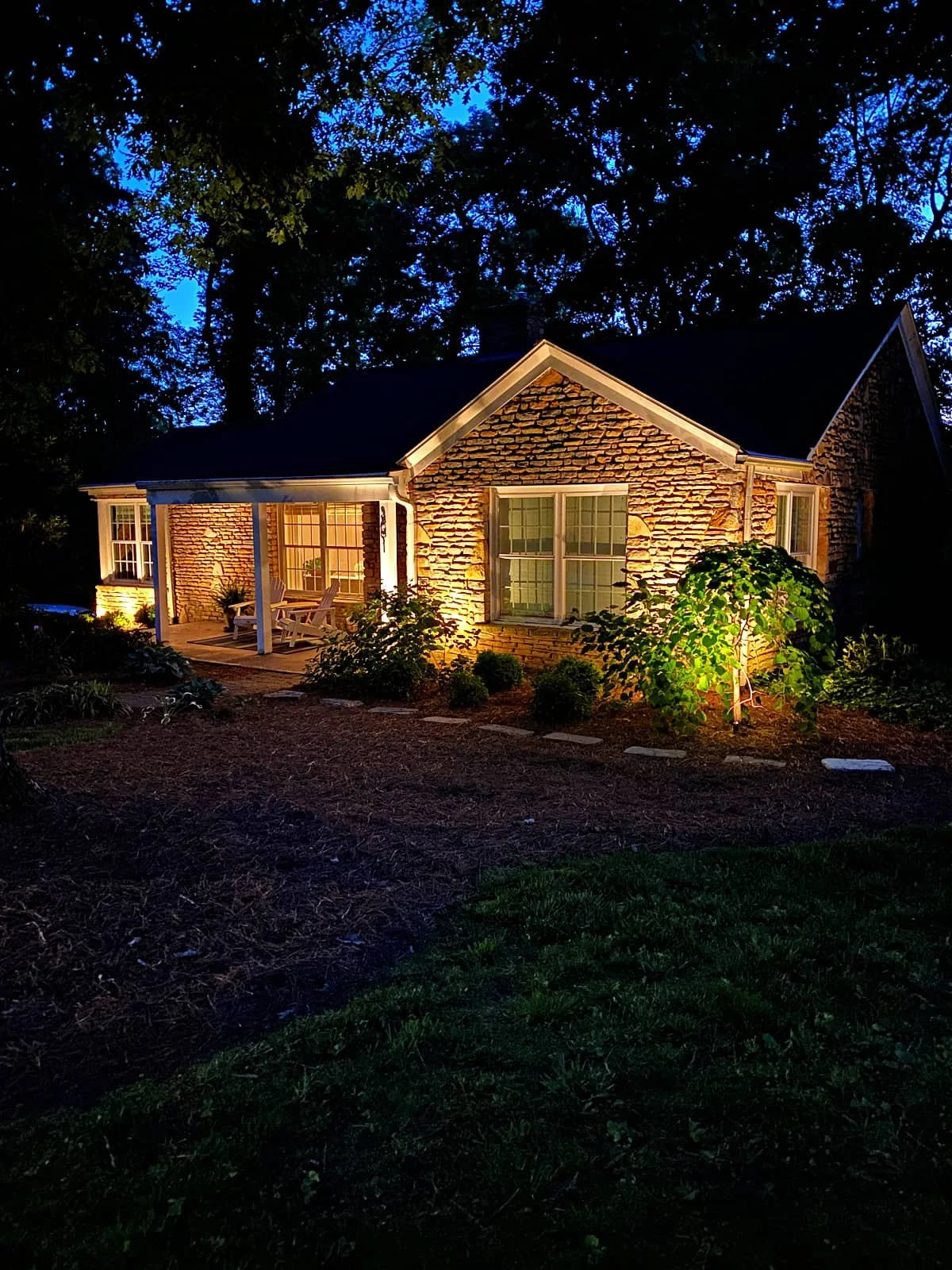 A brick house at night with exterior lighting illuminating the front porch and garden, surrounded by trees.